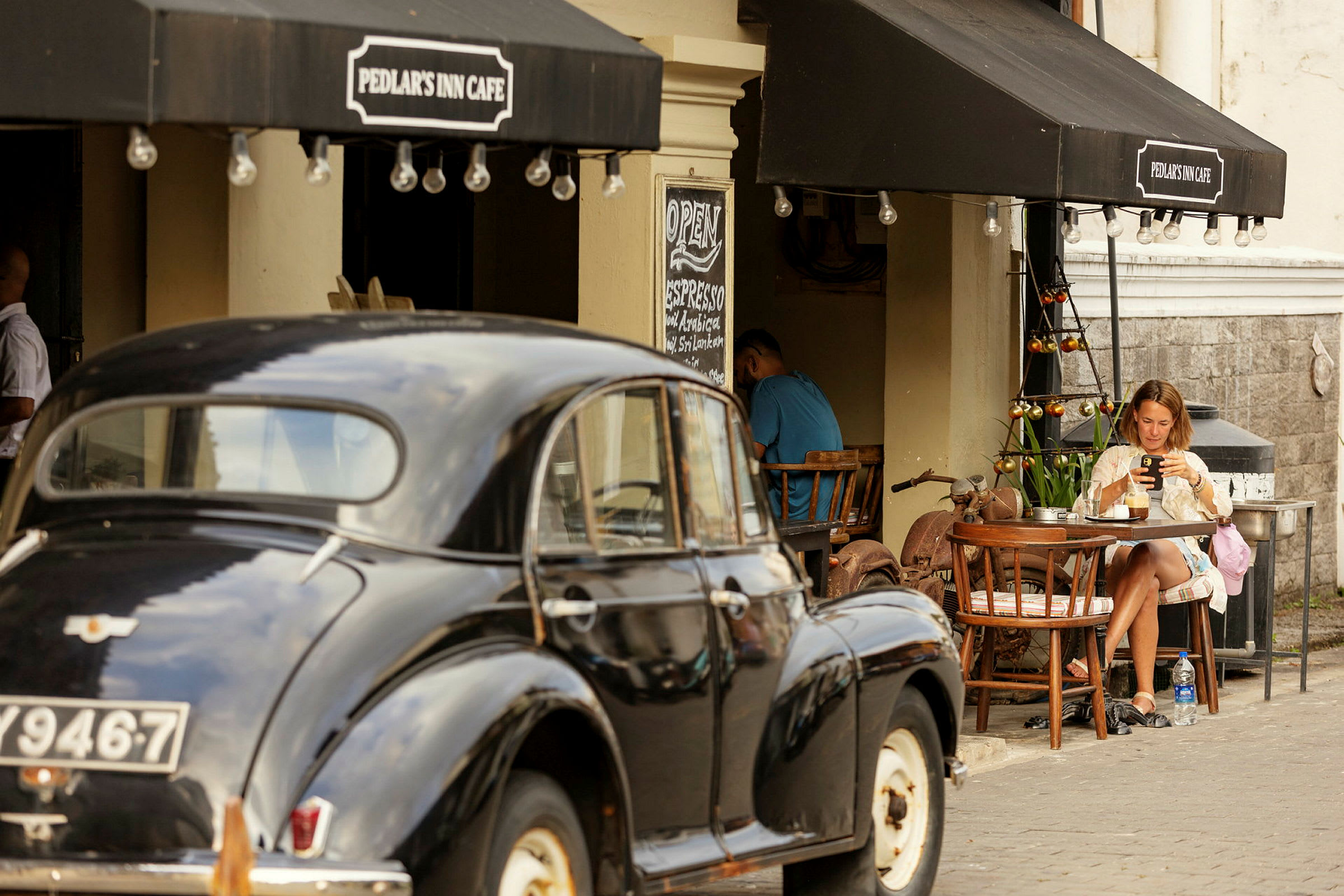 Billede af et udendørs caféområde med en parkeret klassisk bil i forgrunden og en kvinde, der sidder ved et bord. (AI-genereret tekst)