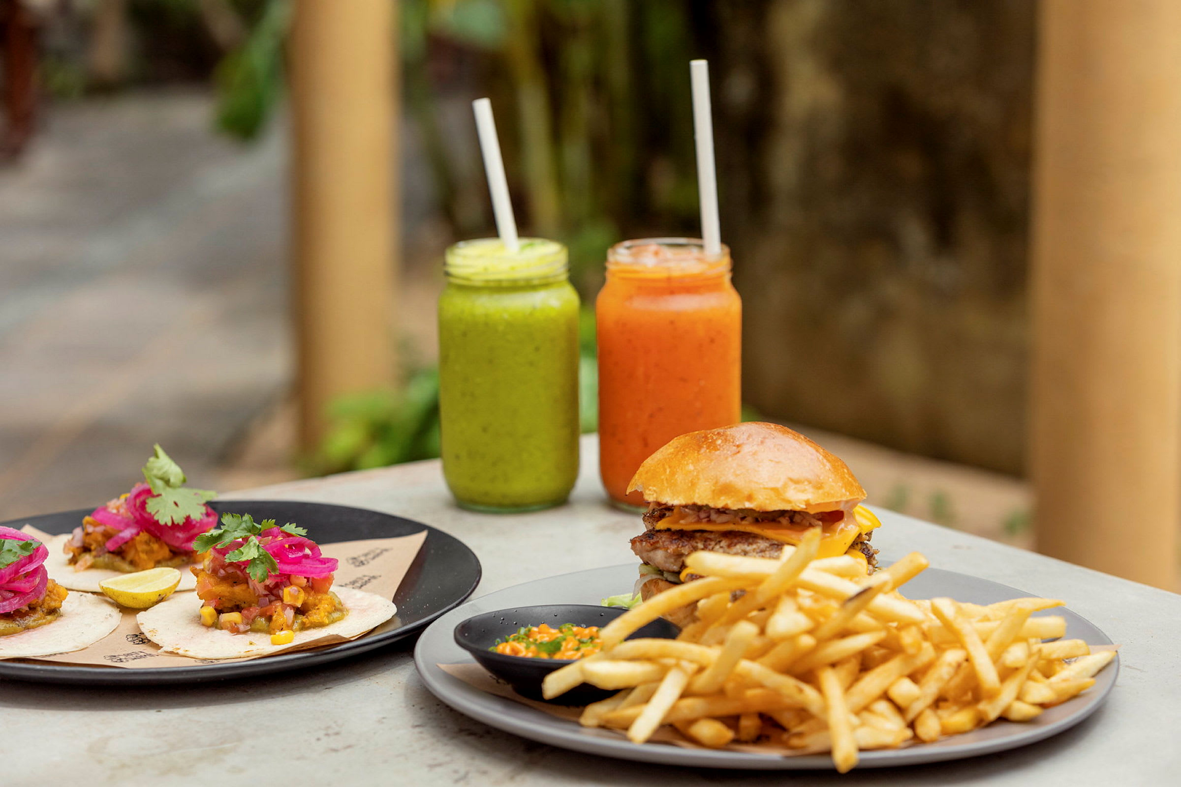 På bordet står en tallerken med tacos, en med burger og pommes fritter samt to glas med farverige drikke. (AI-genereret tekst)