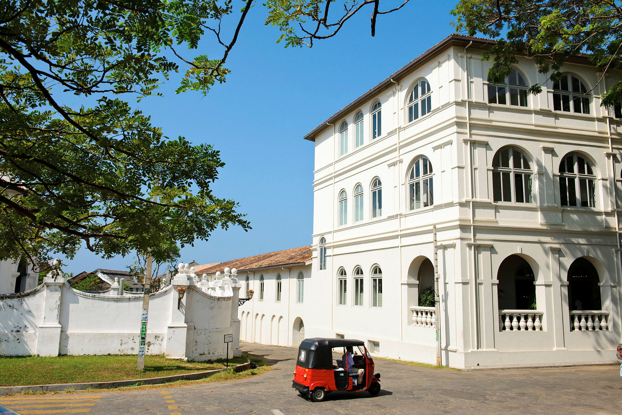 Et hvidt kolonialt bygningskompleks med buede vinduer og detaljer. Foran bygningen er en rød tuk-tuk parkeret, omgivet af grønne områder under en klar blå himmel. (AI-genereret tekst)