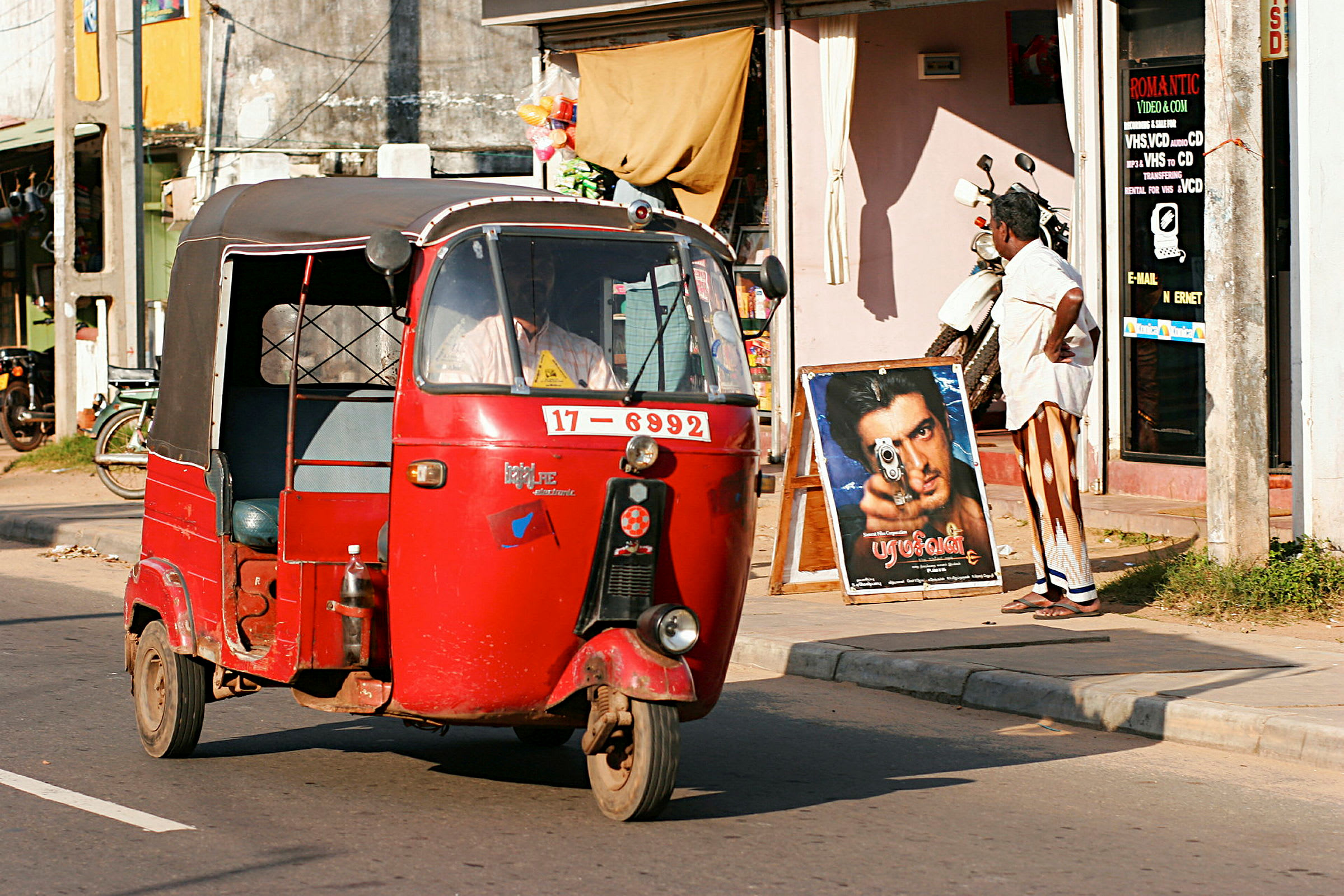 En rød tuk-tuk kører forbi en butik på en solrig gade. (AI-genereret tekst)