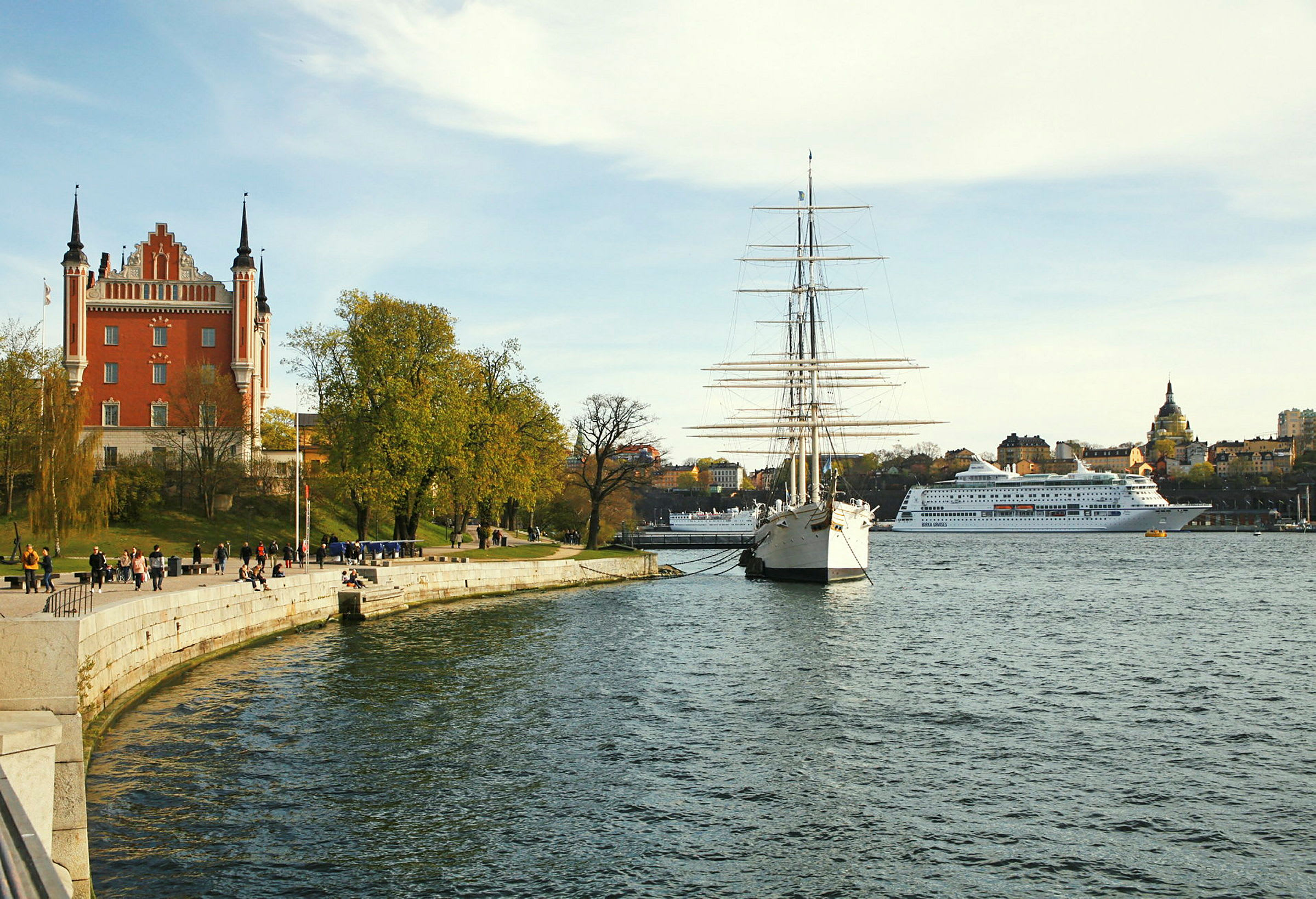 Et rødt hus med tårne ved en kaj, et sejlskib og et krydstogtskib ses ved vandet. (AI-genereret tekst)