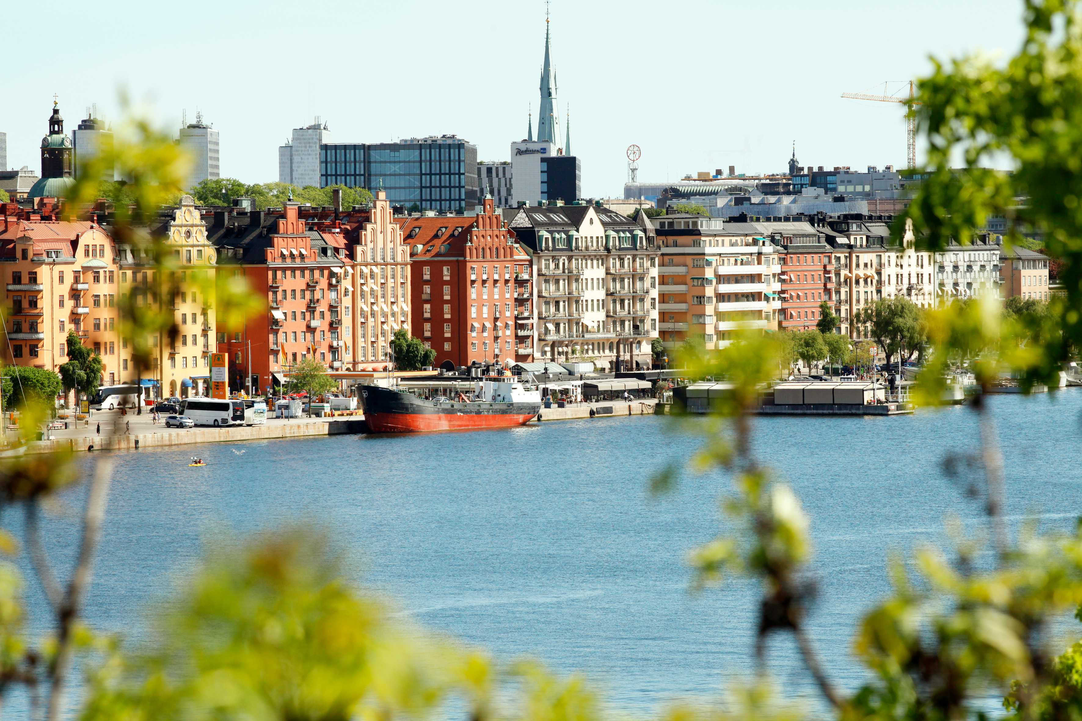 Udsigt over en kystlinje med bygninger og både ved vandet, delvist indrammet af grønt. (AI-genereret tekst)
