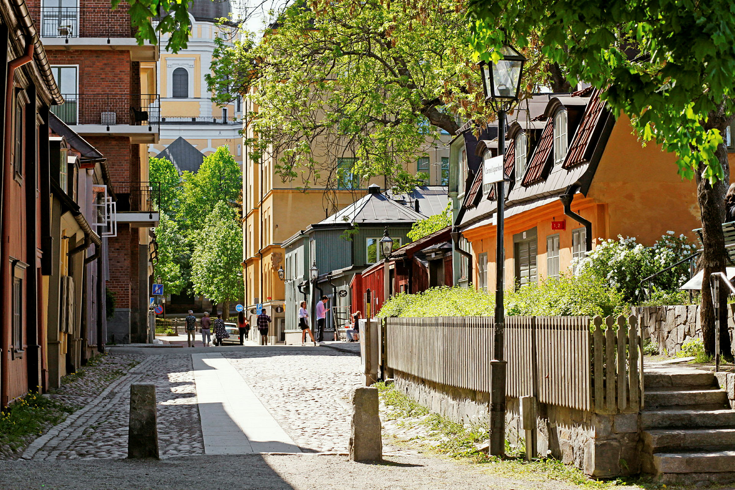 En brostensbelagt gade omkranset af gamle huse, omgivet af grønt og træer. (AI-genereret tekst)