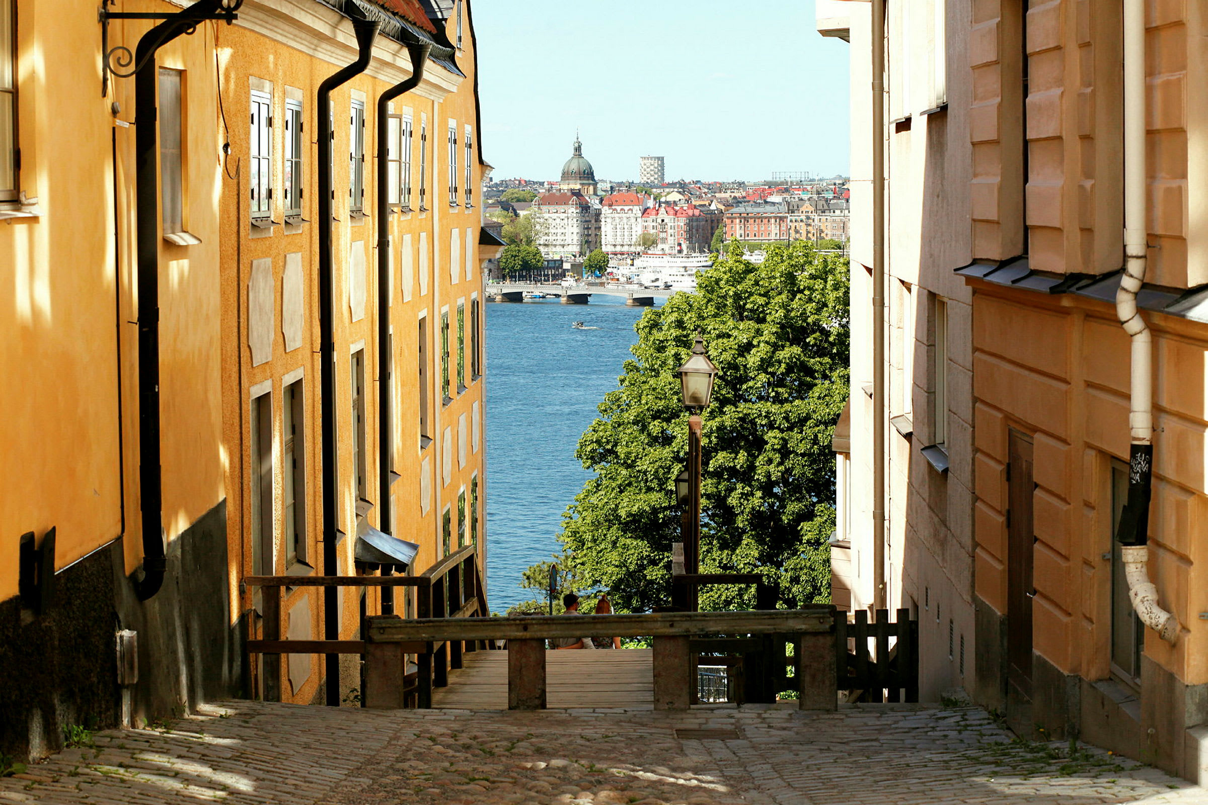 Et gadebillede med orangefarvede bygninger på hver side, som skråner ned mod vand og en by i baggrunden. (AI-genereret tekst)
