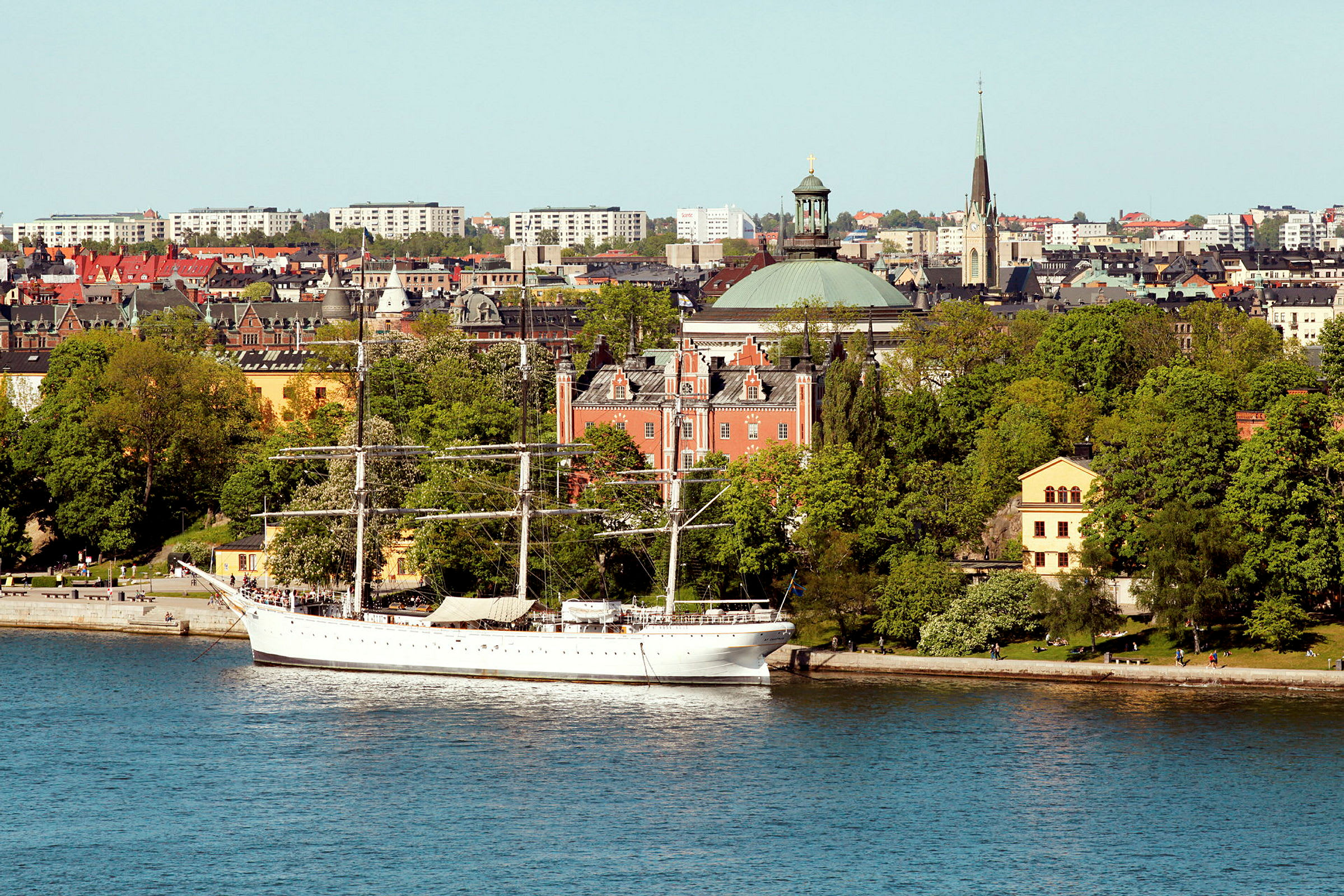 Et hvidt sejlskib ligger for anker ved en kaj i en by ved vandet. Baggrunden udgøres af træer, ældre bygninger og andre bystrukturer. (AI-genereret tekst)