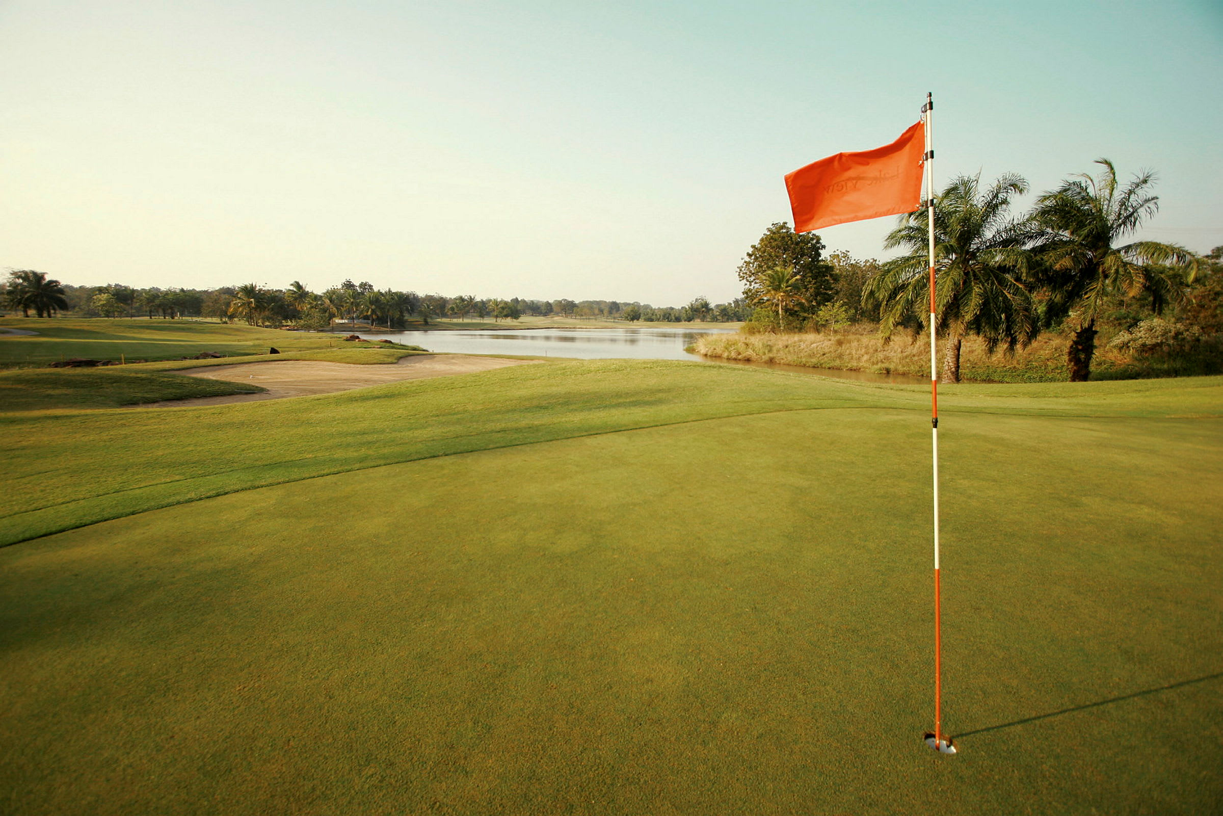 Golfbane med flag ved greenen nær en sø omgivet af palmer. (AI-genereret tekst)