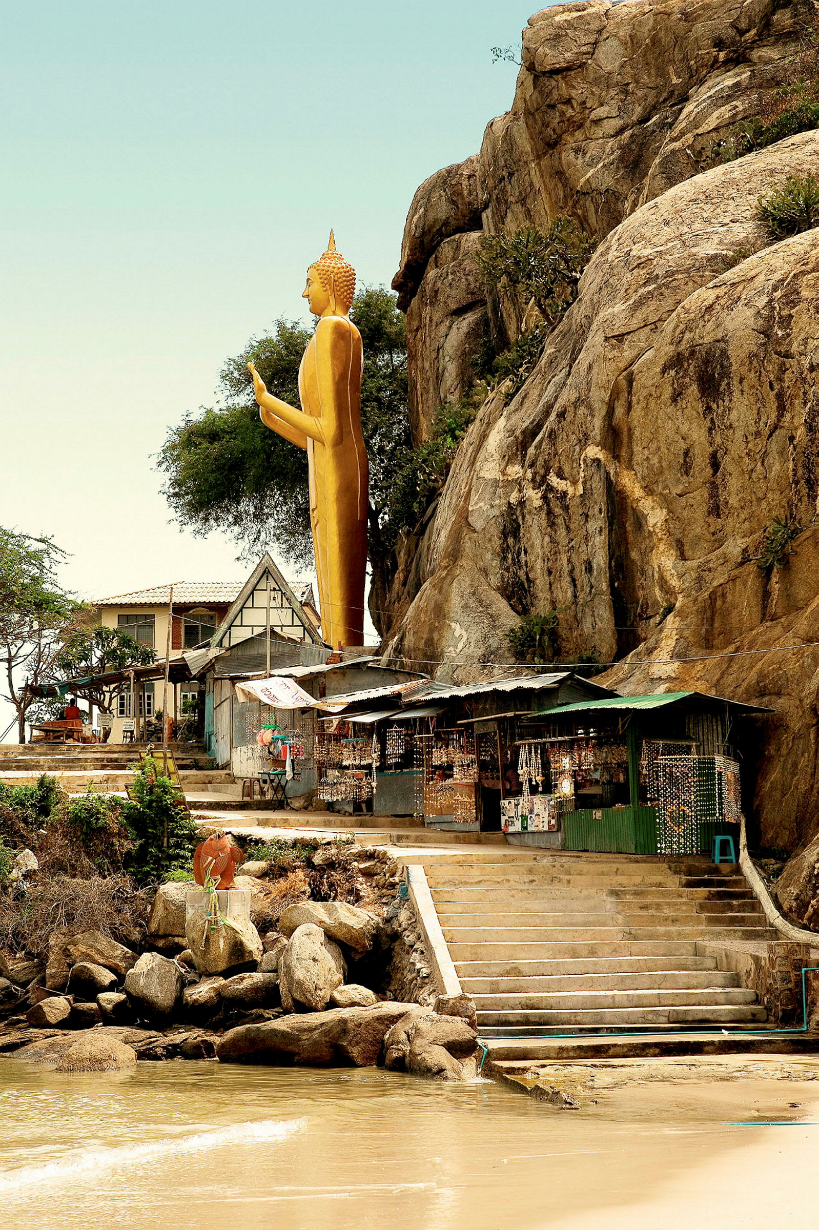 En gylden buddhastatue står ved en klippe over et lille marked og trapper, der fører ned til stranden. (AI-genereret tekst)