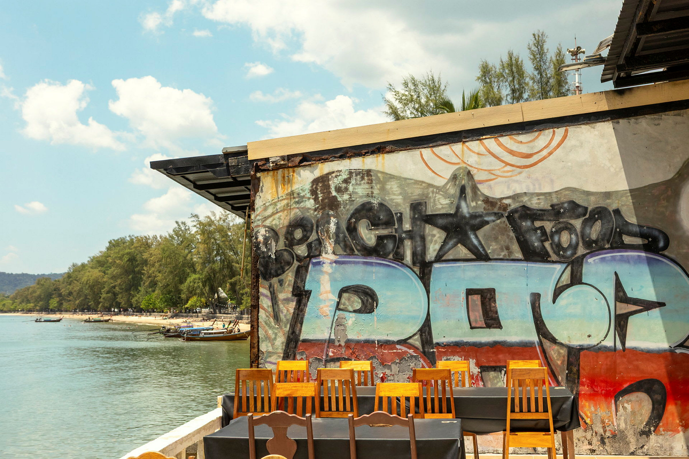 En udsigt over en strand med vand, træer og både. Foran i billedet ses en væg med graffiti samt stole og borde. (AI-genereret tekst)