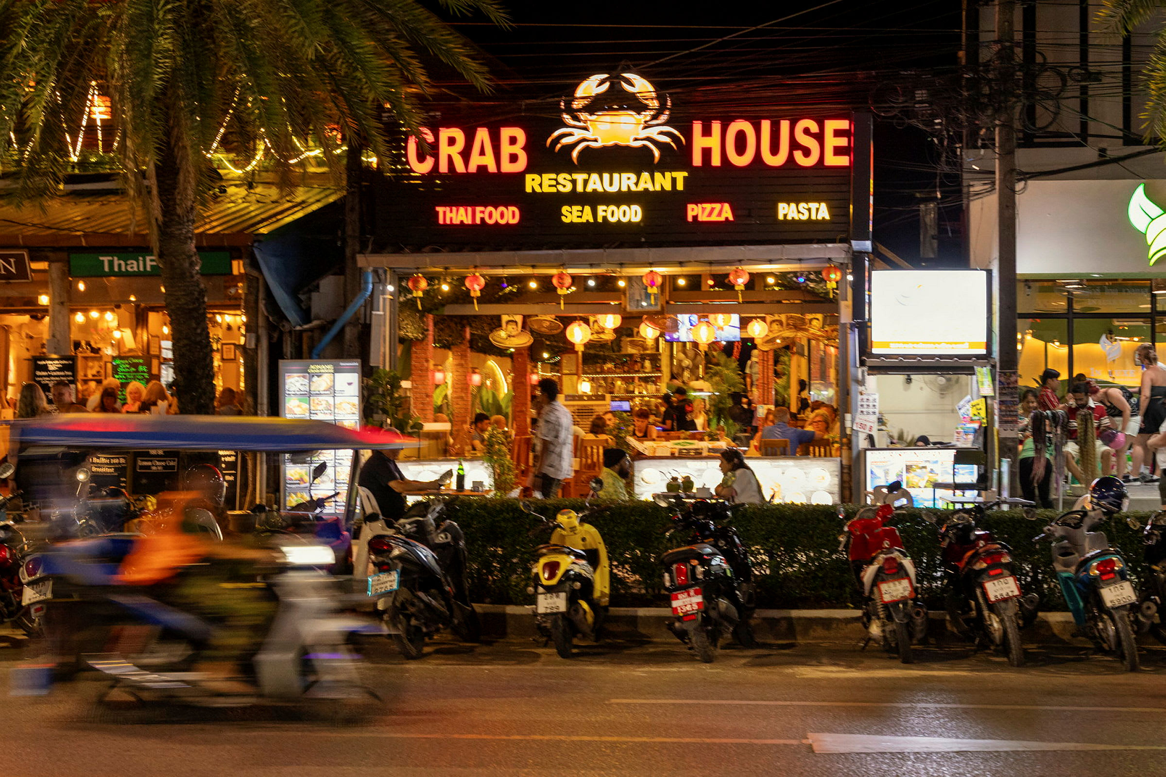 Billede af en livlig gade med restauranten Crab House i fokus. Flere motorcykler er parkeret foran restauranten, og en motorcykel med en passager kører forbi. (AI-genereret tekst)