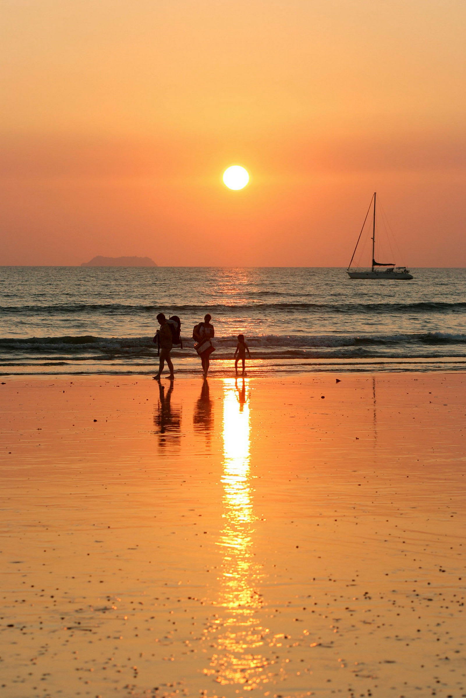 Fire personer går på stranden ved solnedgang, med en sejlbåd synlig på havet. (AI-genereret tekst)