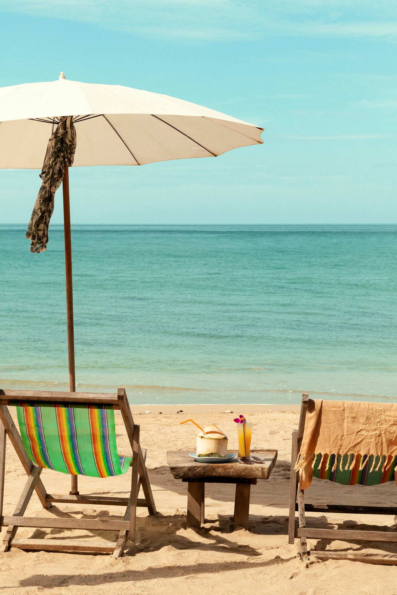 To strandstole, en parasol og et bord med drikkevarer på en sandstrand ved havet. (AI-genereret tekst)