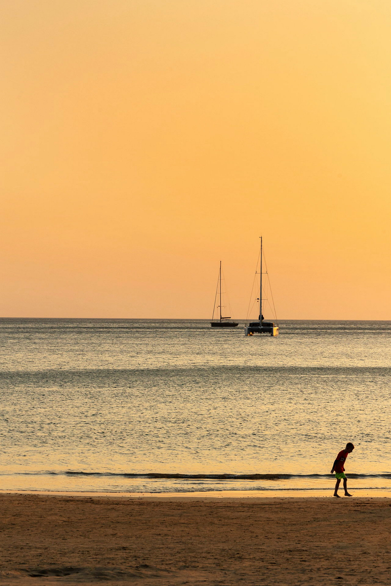 En strand ved havet under en orange solnedgang med to sejlbåde i baggrunden og en person, der går i sandet. (AI-genereret tekst)
