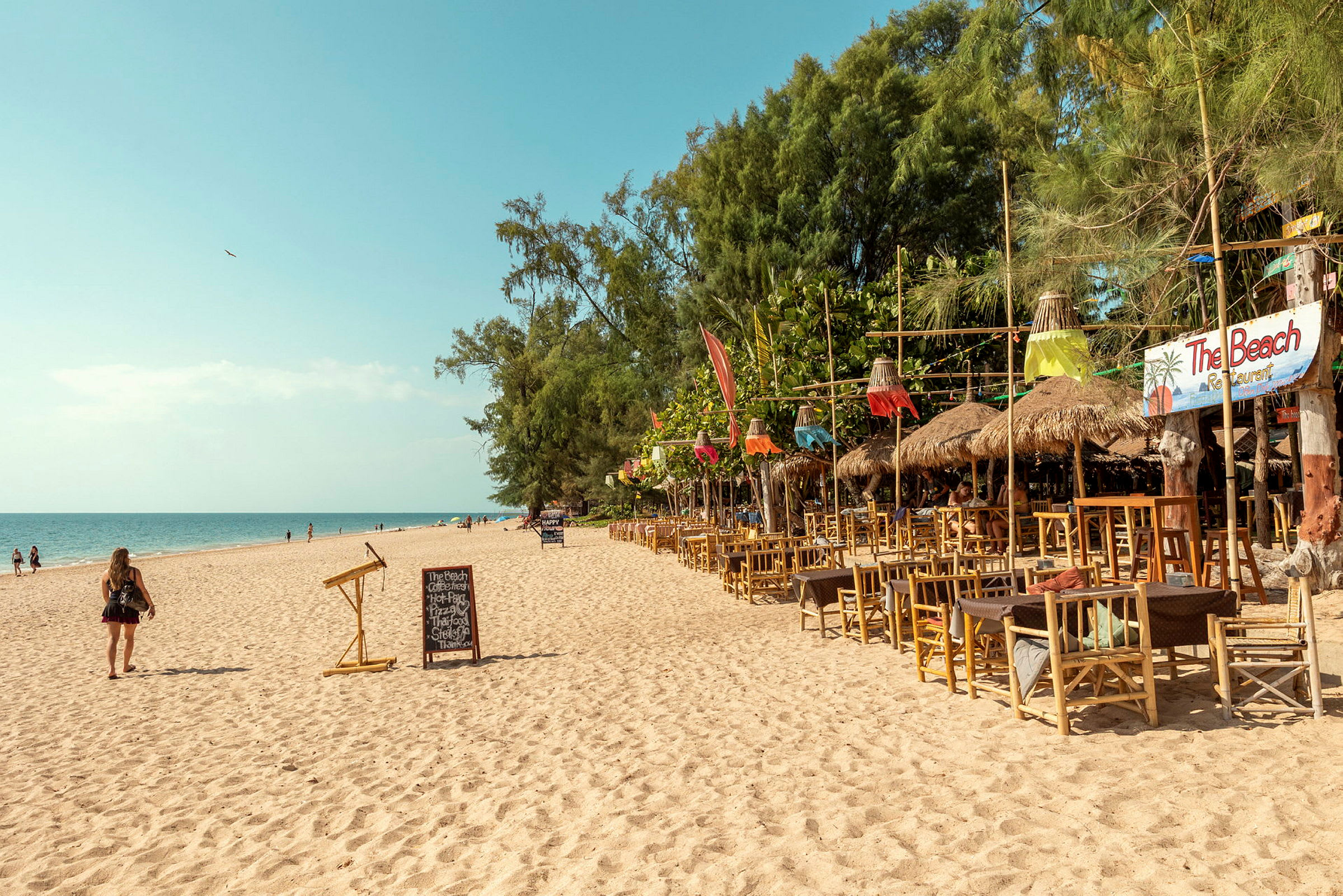 En sandstrand med en række træmøblerede restaurantområder med dekorationer til højre og mennesker, der går ved havet. (AI-genereret tekst)