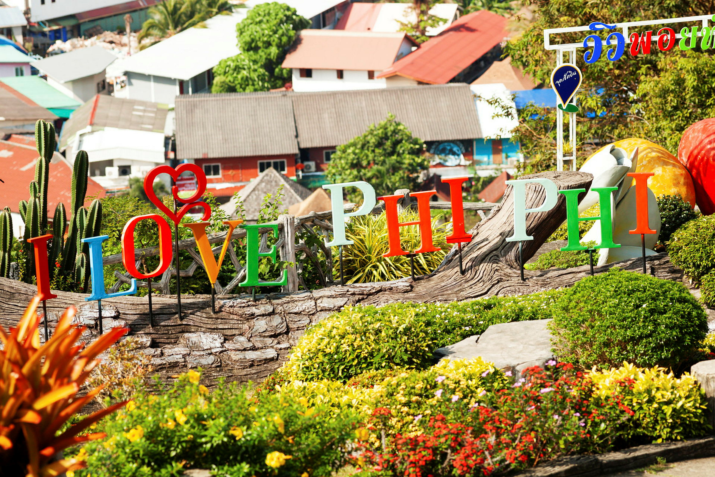 Farverige bogstaver, der staver "I Love Phi Phi", er placeret i et haveområde med buske og blomster. (AI-genereret tekst)