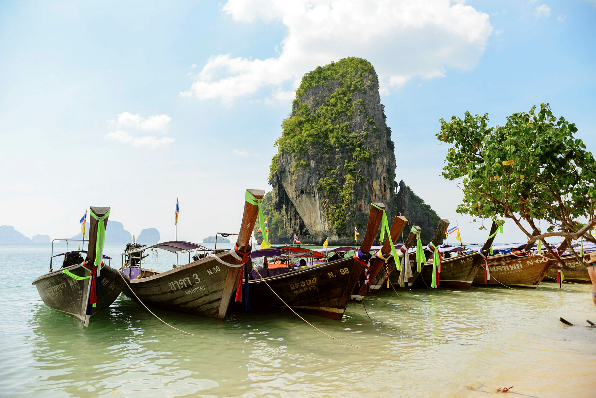 Traditionelle thailandske både fortøjet ved en strand med en kalkstensklippe i baggrunden. (AI-genereret tekst)