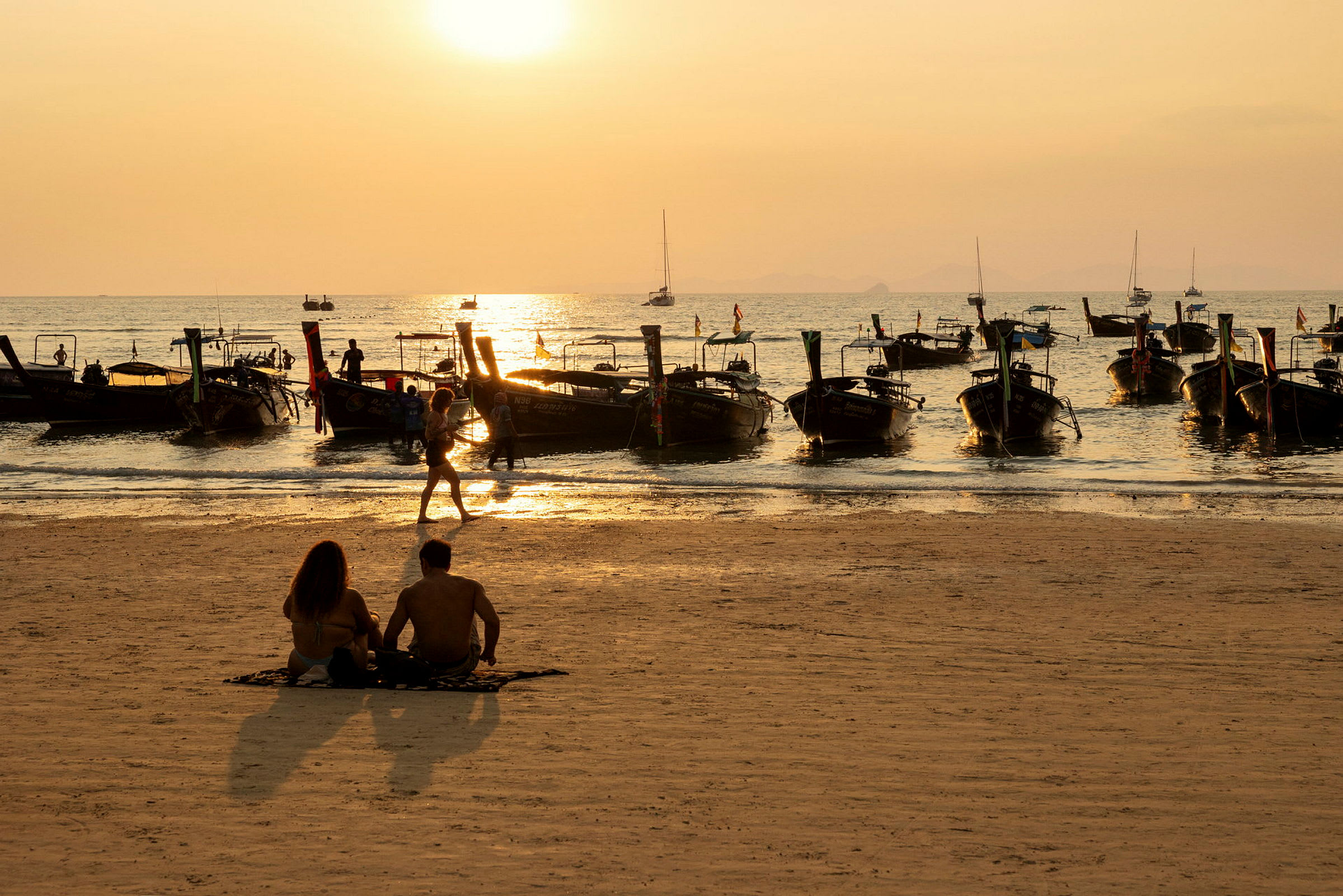 En strand med mennesker der sidder og går, og både fortøjet tæt på stranden ved solnedgang. (AI-genereret tekst)