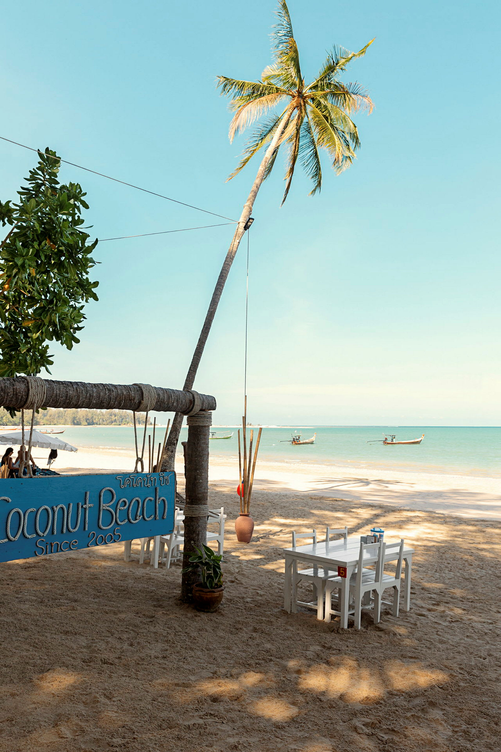 En strand med palmer, borde og stole samt et skilt, der markerer Coconut Beach. (AI-genereret tekst)