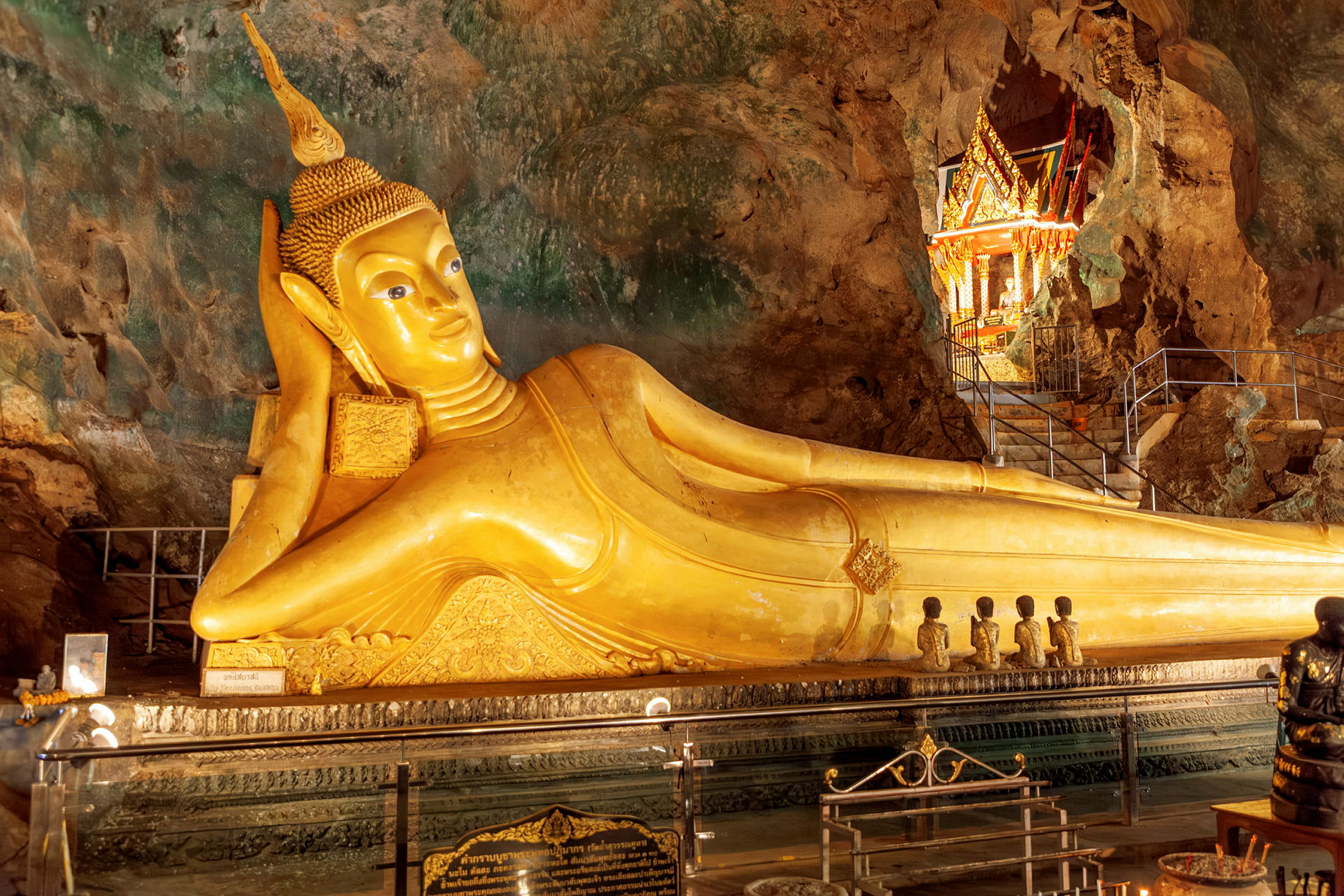 En gylden liggende Buddha-statue inde i en klippehule omgivet af et smukt dekoreret område med detaljerede udsmykninger. (AI-genereret tekst)