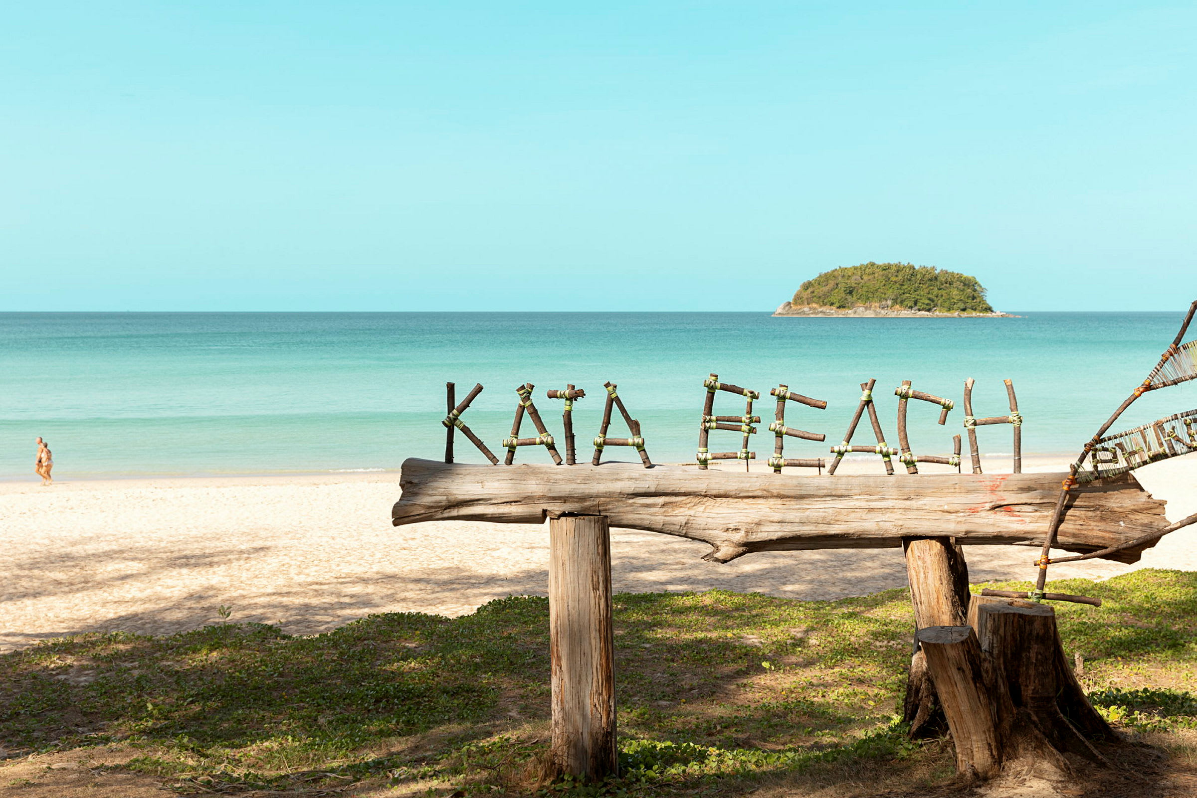 Træskilt med teksten 'Kata Beach' på en sandstrand med havet i baggrunden. (AI-genereret tekst)