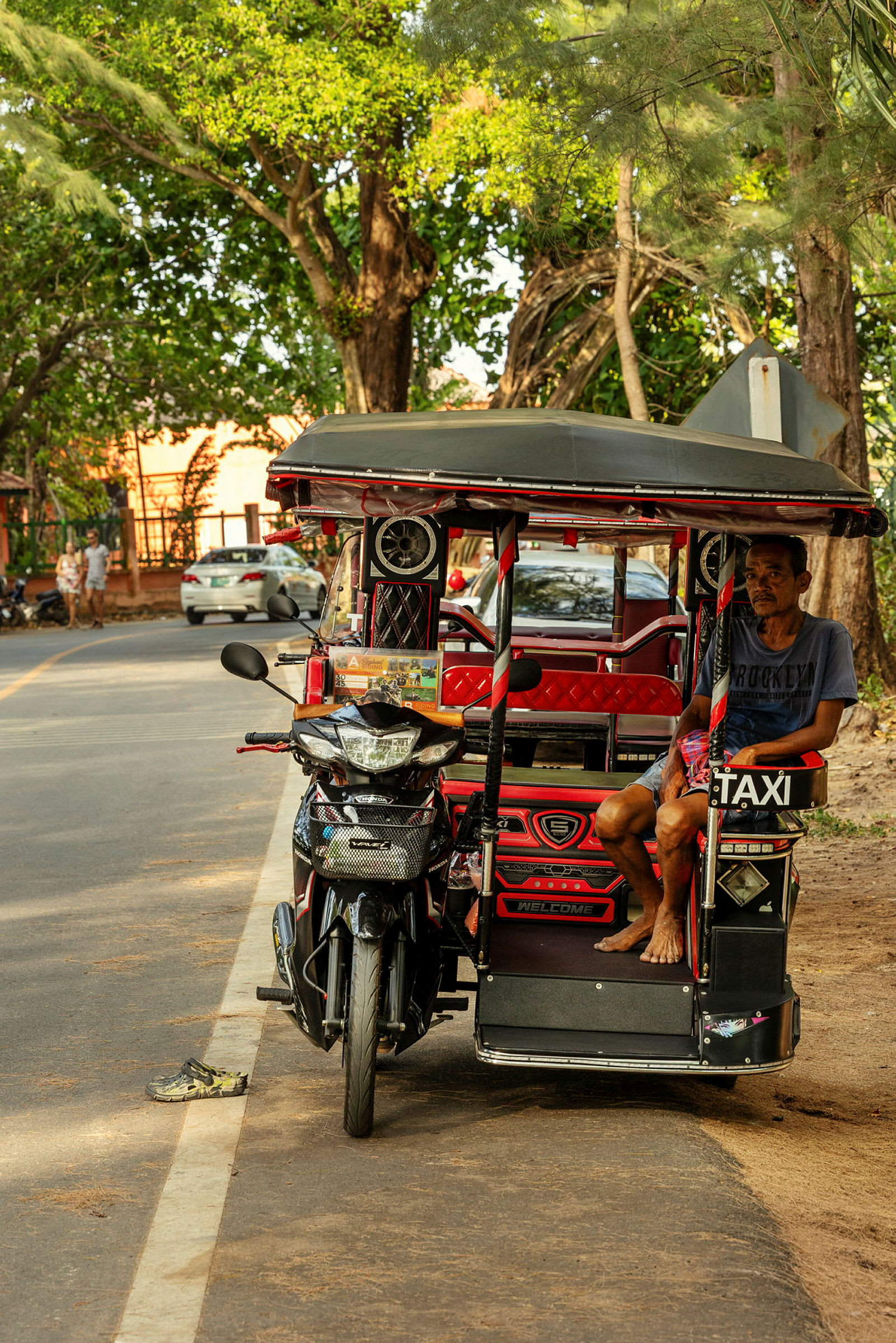 En tuk-tuk står parkeret ved siden af en vej. En mand sidder i køretøjet, omgivet af træer og nogle parkerede biler. (AI-genereret tekst)