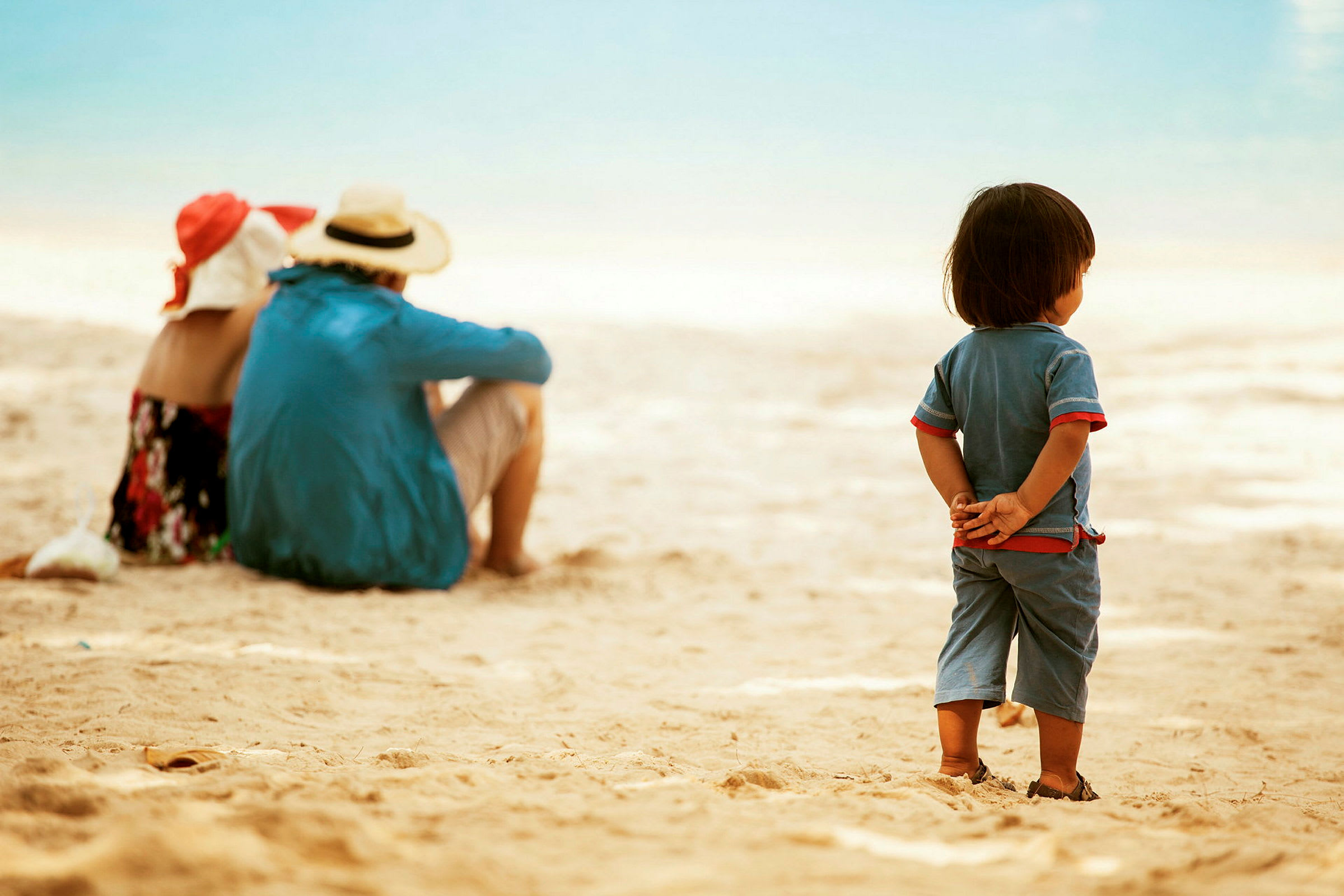 Et barn står på stranden med hænderne bag ryggen, mens to voksne sidder længere væk. (AI-genereret tekst)