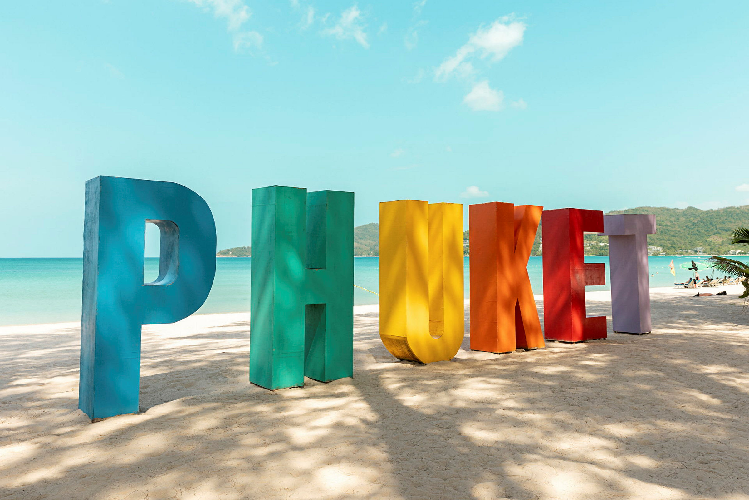 Farverige bogstaver former ordet 'Phuket' på en sandstrand, med hav og himmel i baggrunden. (AI-genereret tekst)