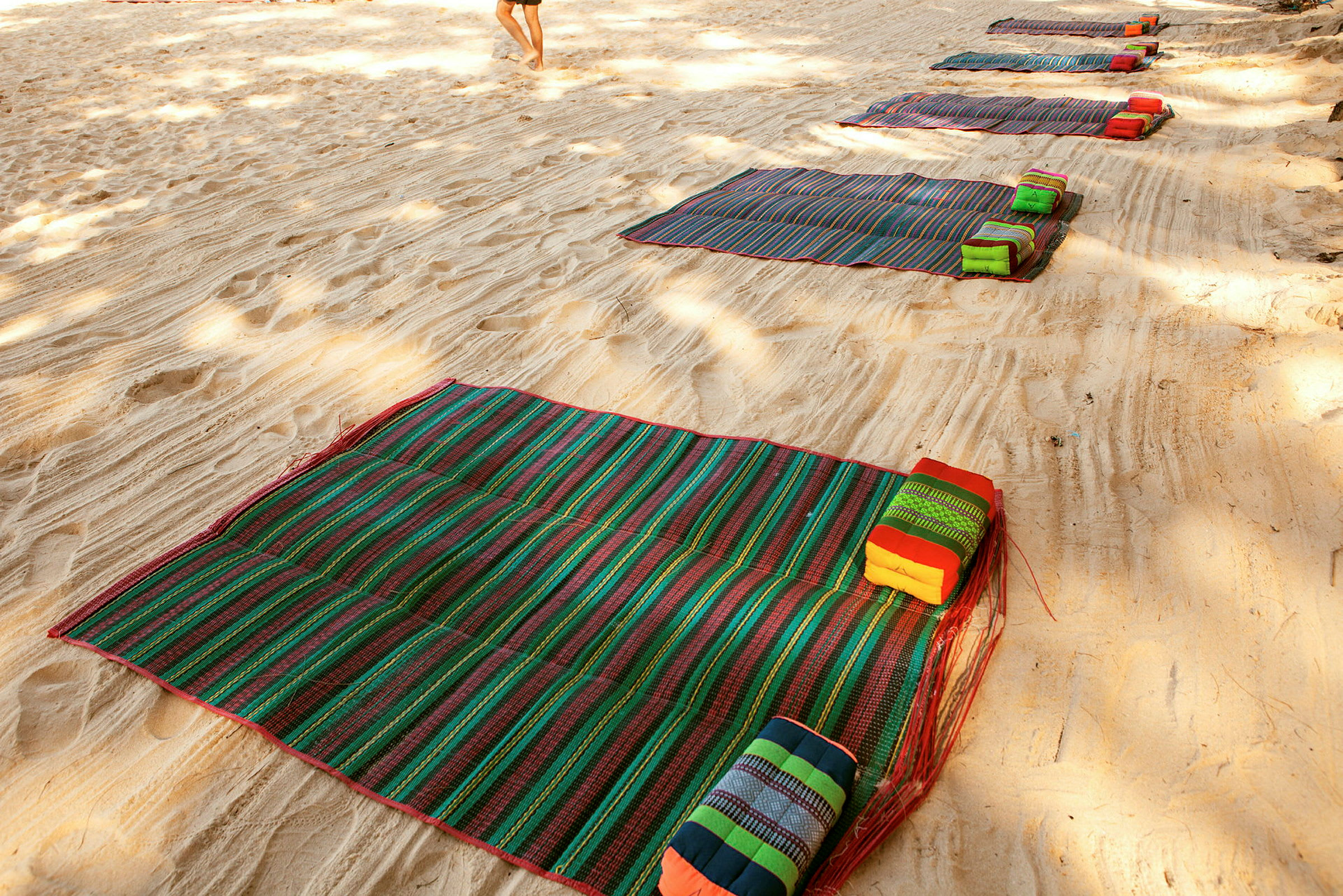 Flere farverige strandmåtter med puder placeret på sandstrand. (AI-genereret tekst)