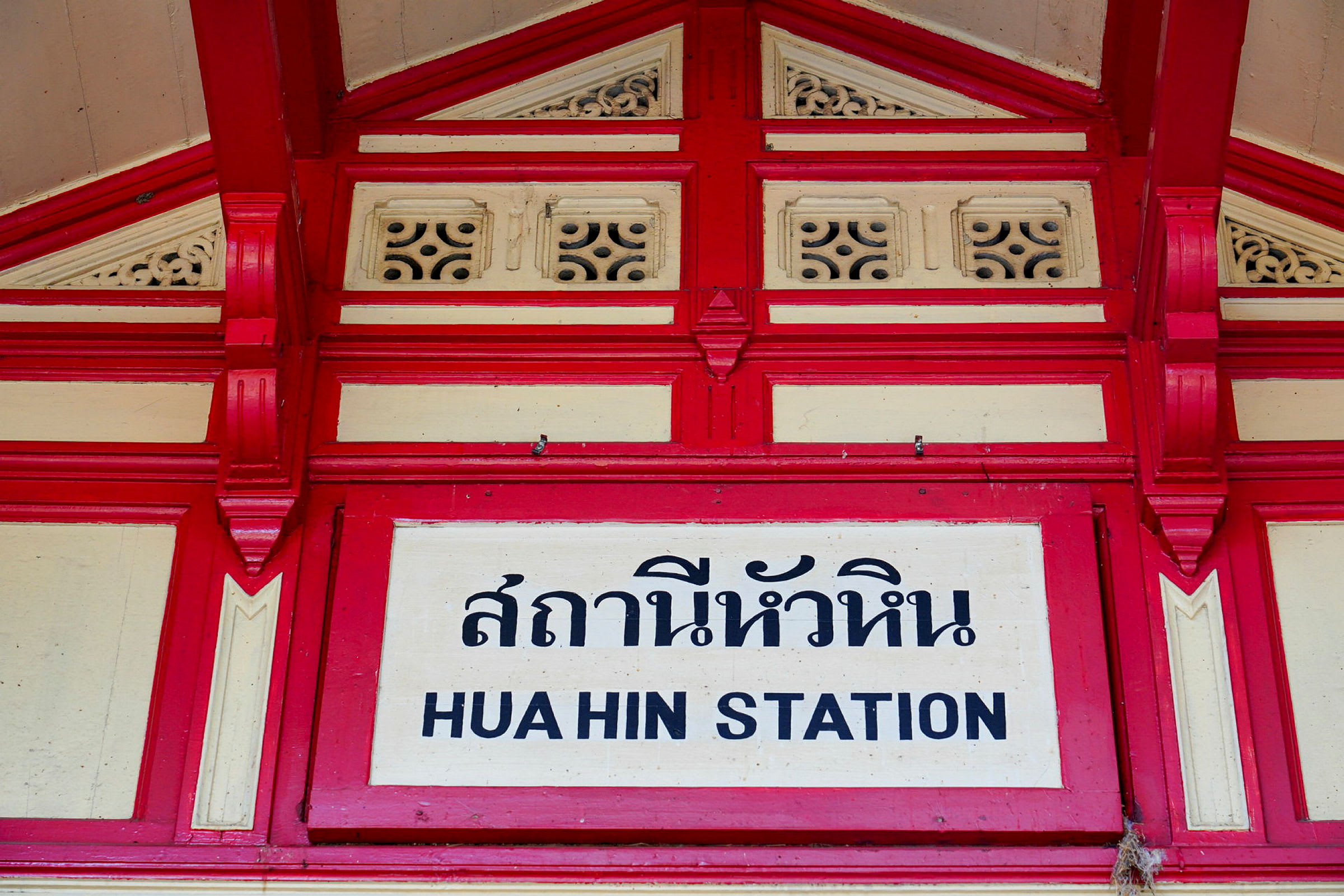 Skilt med teksten 'Hua Hin station' med thailandsk skrift og dekorativ rød og hvid ramme. (AI-genereret tekst)