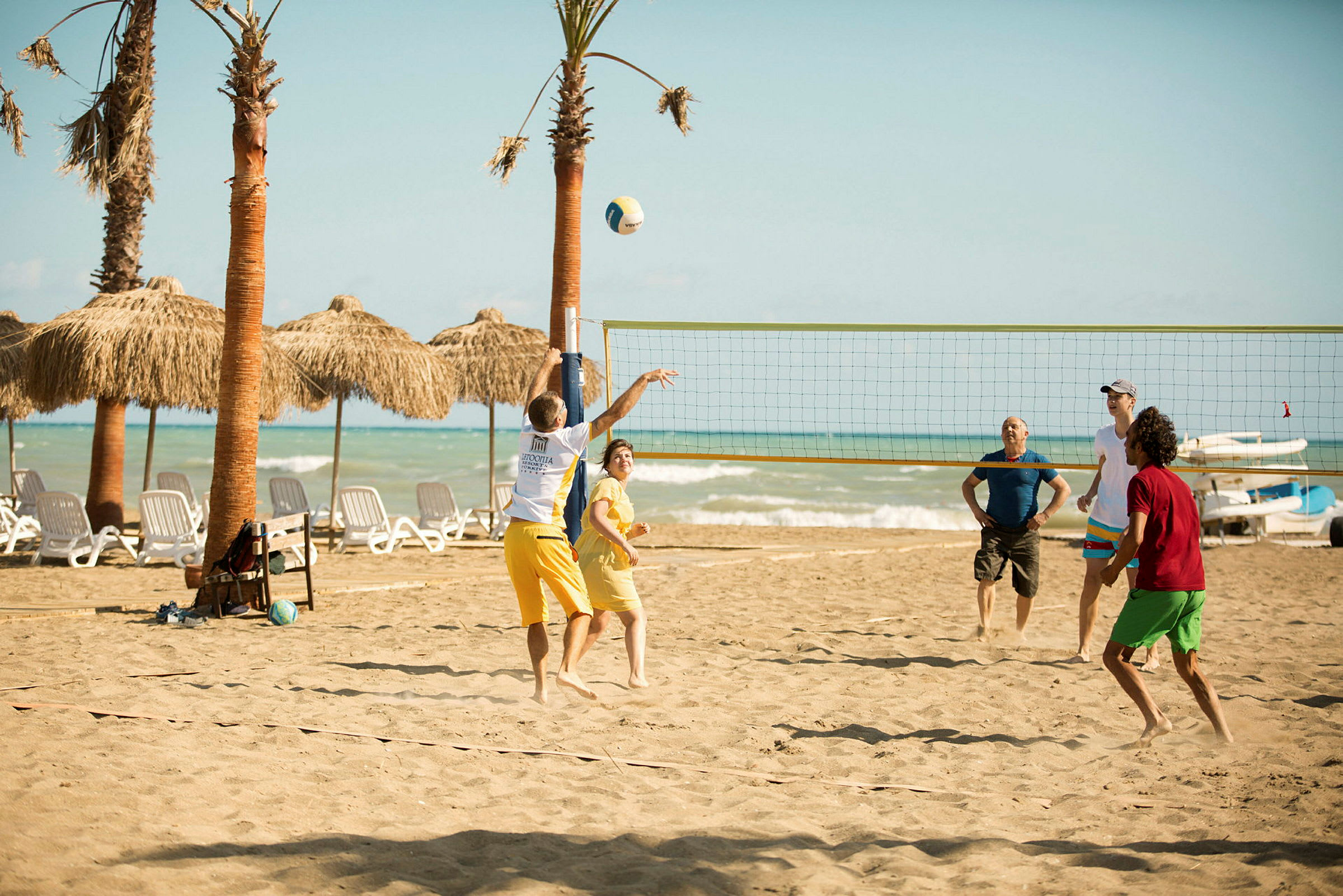 Personer spiller volleyball på en sandstrand med palmer og hav i baggrunden. (AI-genereret tekst)