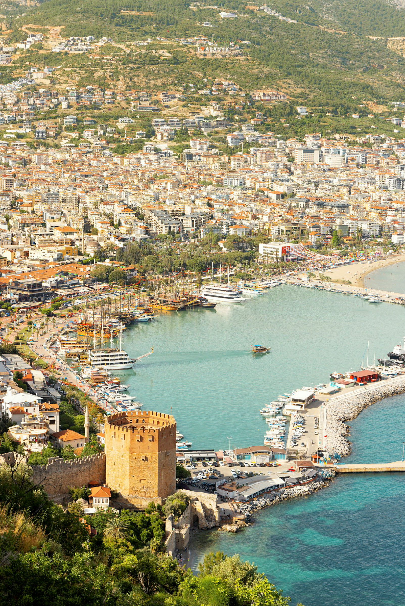 Udsigt over havn og byen Alanya med en historisk rød bygning i forgrunden. (AI-genereret tekst)