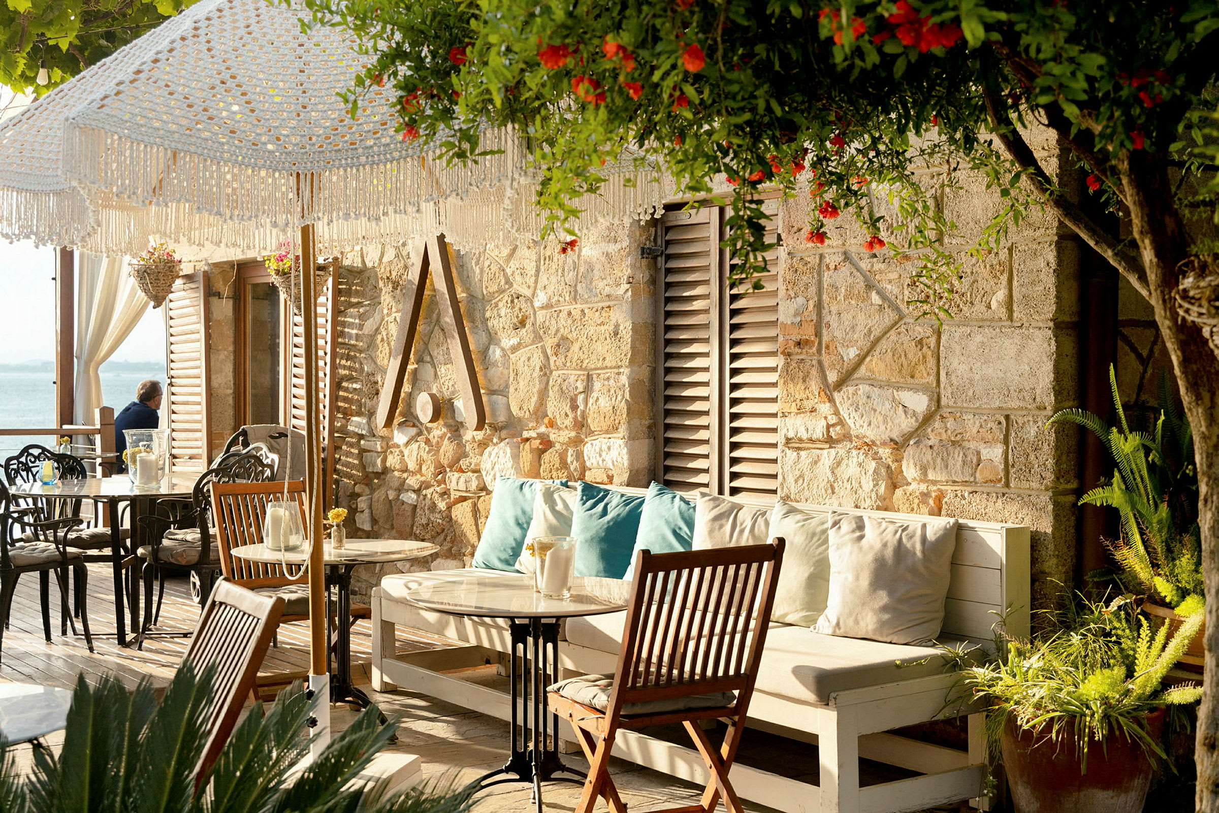 Billede af en terrasse med siddepladser, borde og planter ved en murstensvæg. Solskin med lysdekorationer og hav i baggrunden. (AI-genereret tekst)