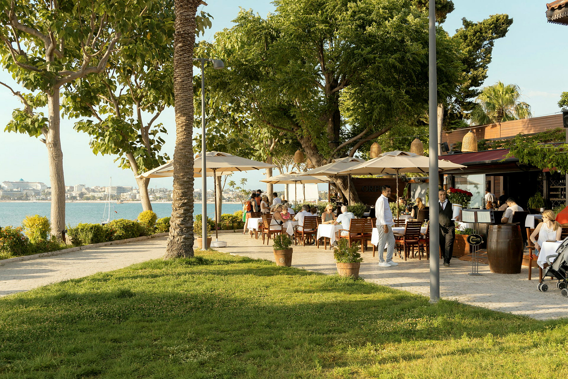 Restaurant med udendørs servering, borde med hvide duge og frodige træer nær vandet. (AI-genereret tekst)