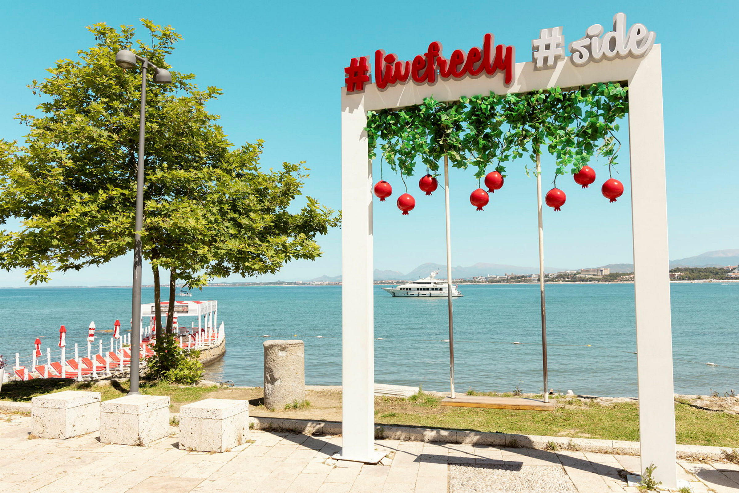 En dekorativ bue med teksten '#livefreely #side', hængende røde dekorationer, ved havet foran en brygge og en yacht i baggrunden. (AI-genereret tekst)