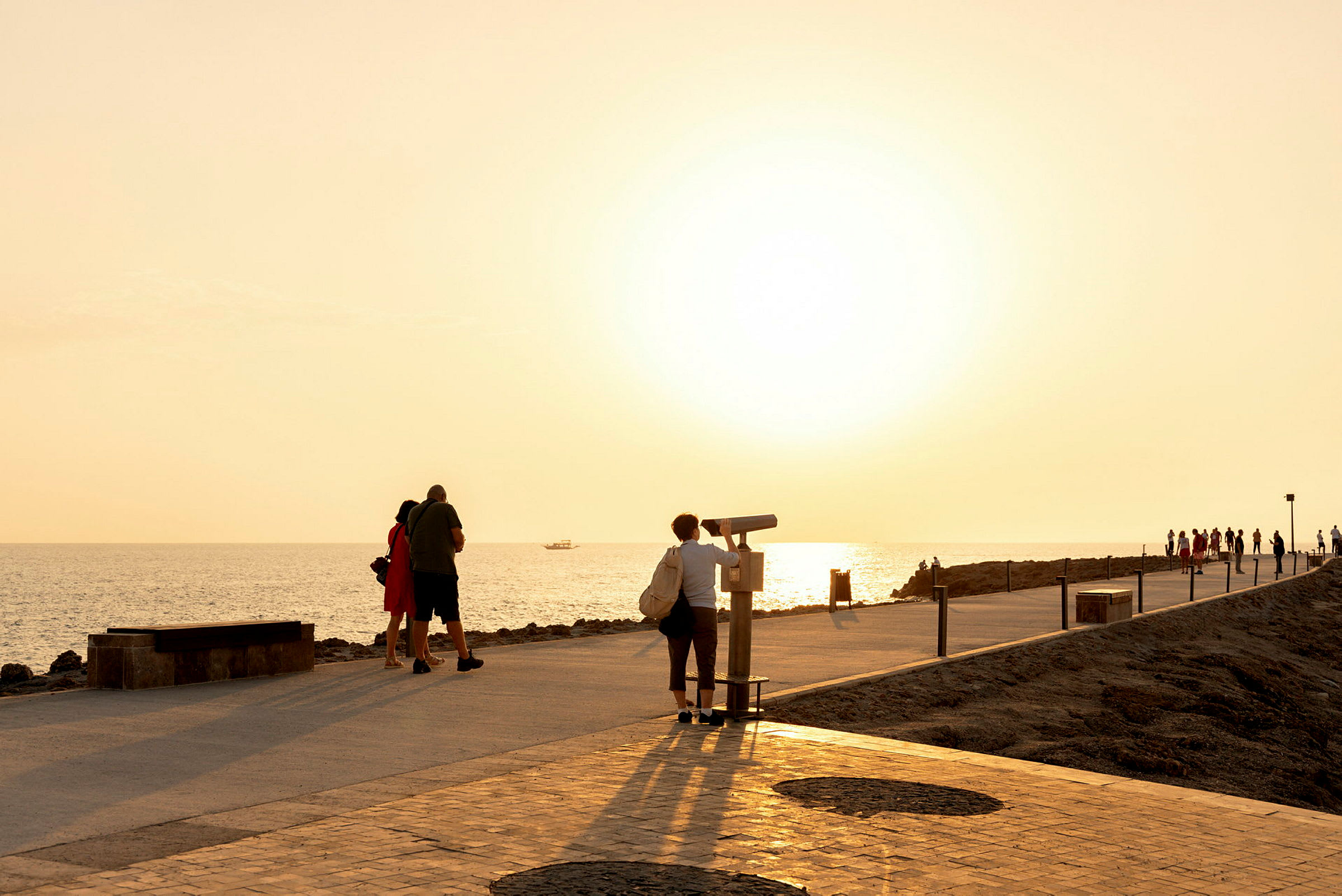 Personer spadserer på en strandpromenade ved havet under solnedgangen. (AI-genereret tekst)