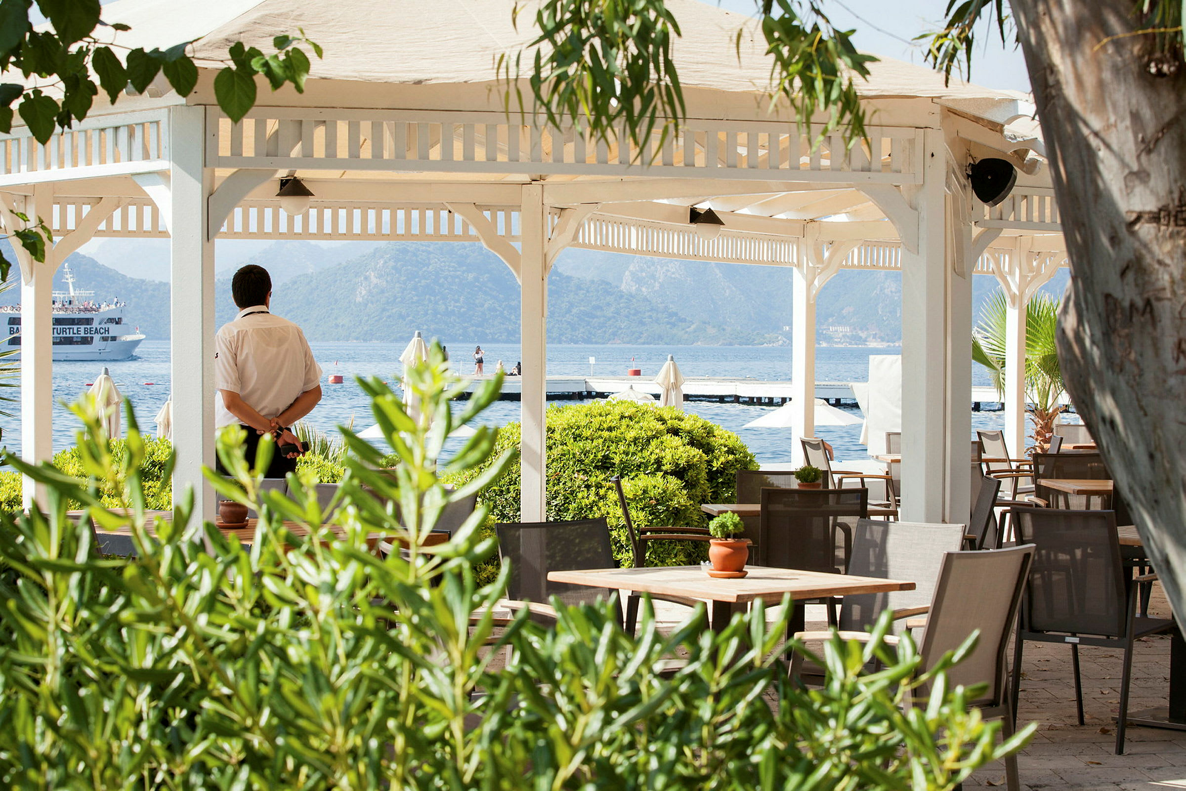 En åben pavillon med borde og stole ved havet, omgivet af grønt. (AI-genereret tekst)