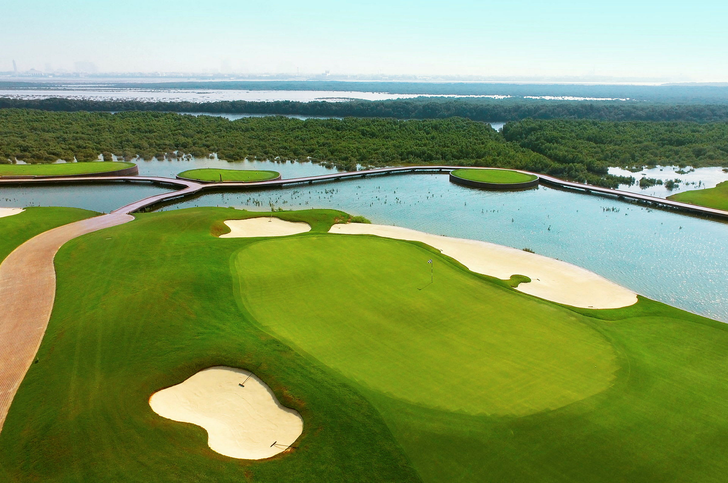 Billedet viser en golfbane med sandbunkere og grønne fairways, omgivet af vand og vegetation. (AI-genereret tekst)