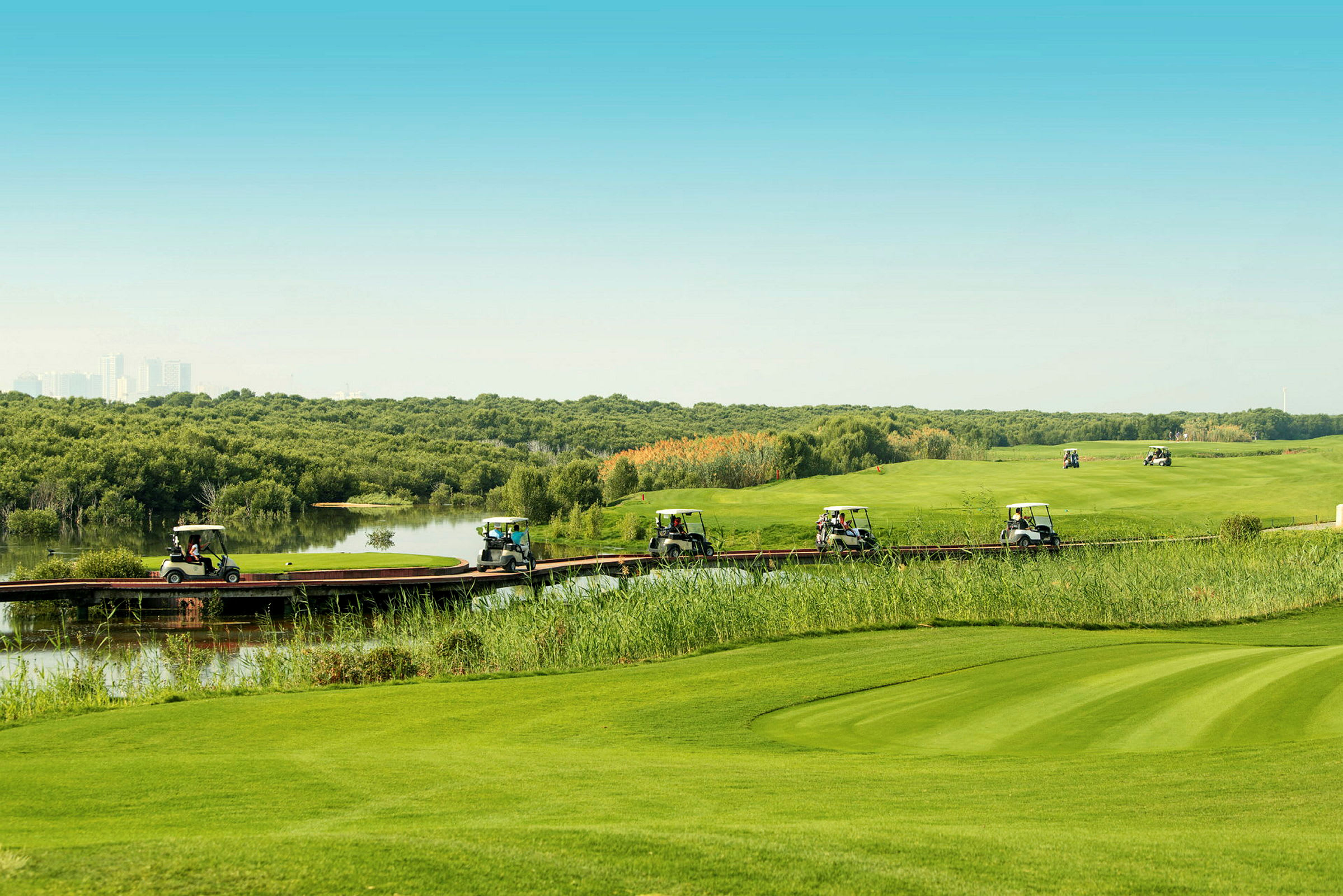 Flere golfbiler kører på en smal træbro, der går gennem et grønt landskab med vand og vegetation. (AI-genereret tekst)