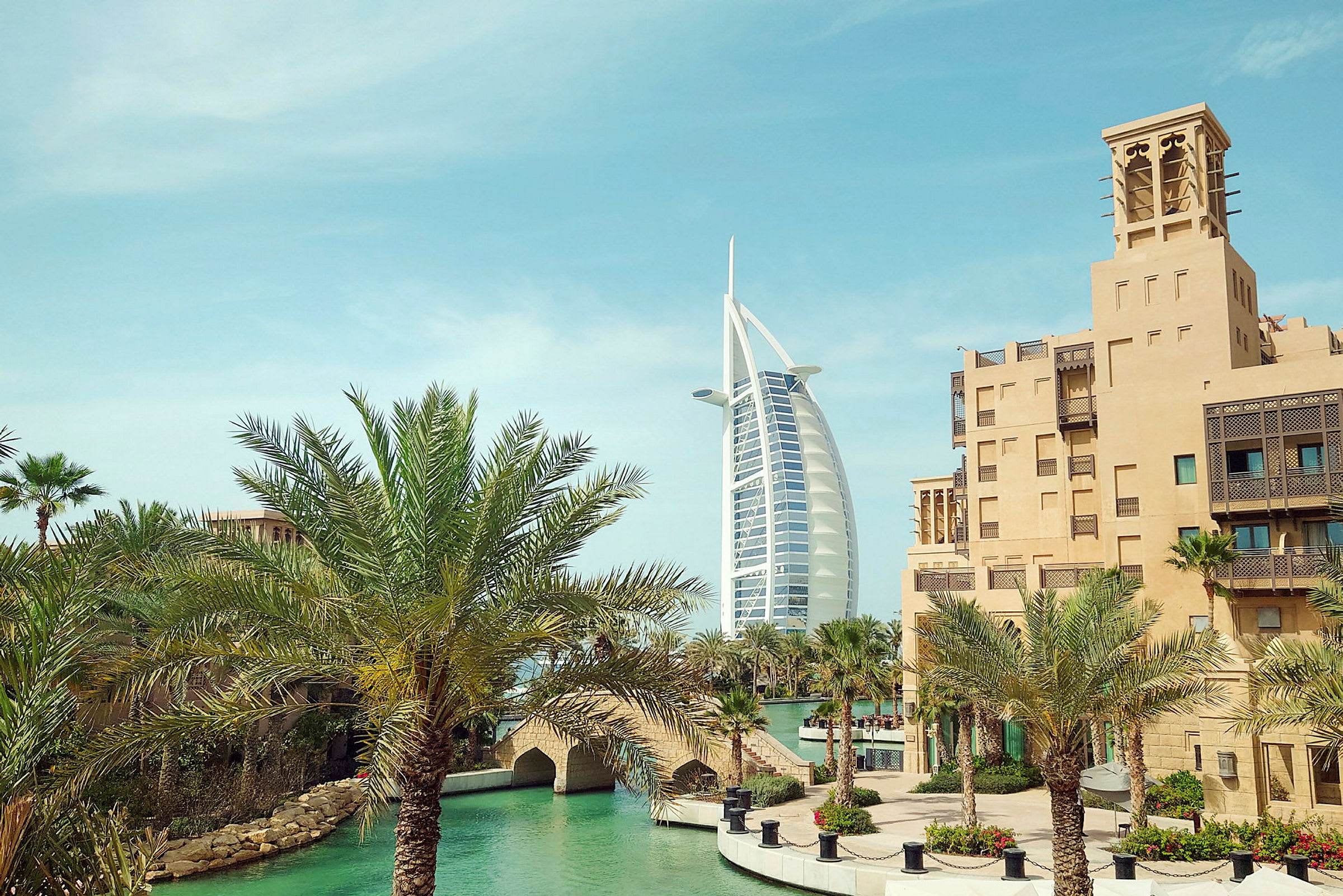 Billede af Burj Al Arab med traditionelle bygninger og palmer i forgrunden. (AI-genereret tekst)