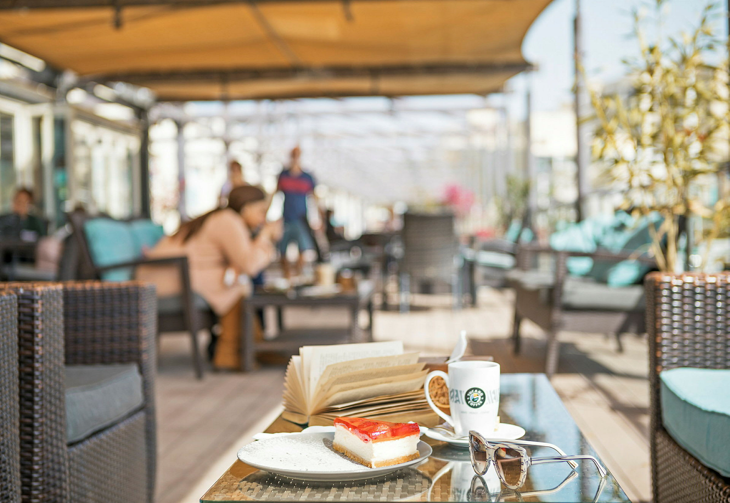 Mad og drikke på et bord med en kaffekop, en bog og en dessert i en udendørs lounge. (AI-genereret tekst)
