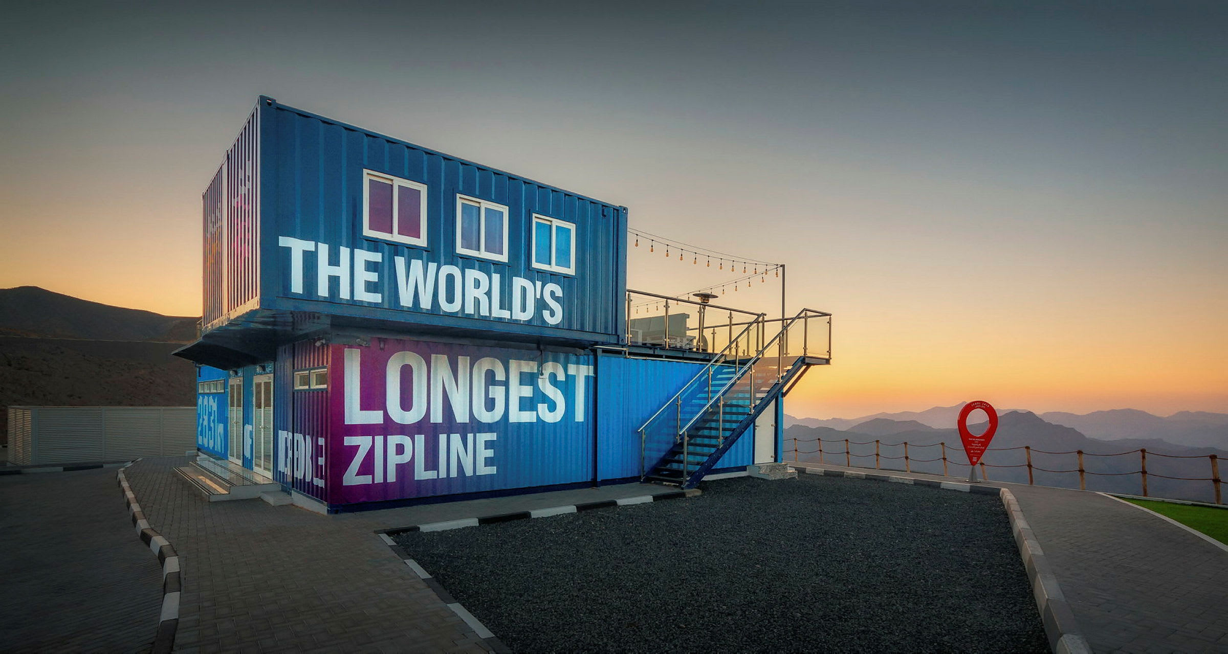 Blå og lilla bygning med teksten 'The World's Longest Zipline' ved solnedgang, omgivet af bjerge. (AI-genereret tekst)