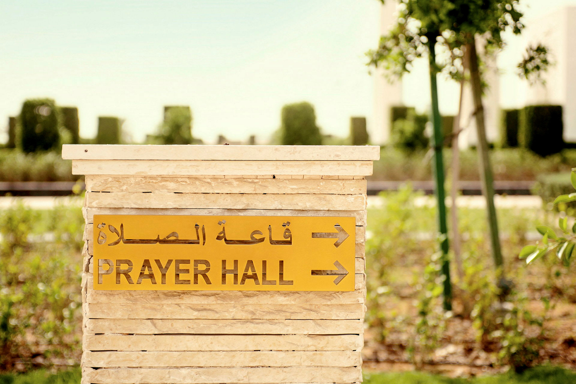 Et skilt med teksten 'Prayer Hall' og arabisk tekst peger mod en bønnehal, omgivet af grønt. (AI-genereret tekst)