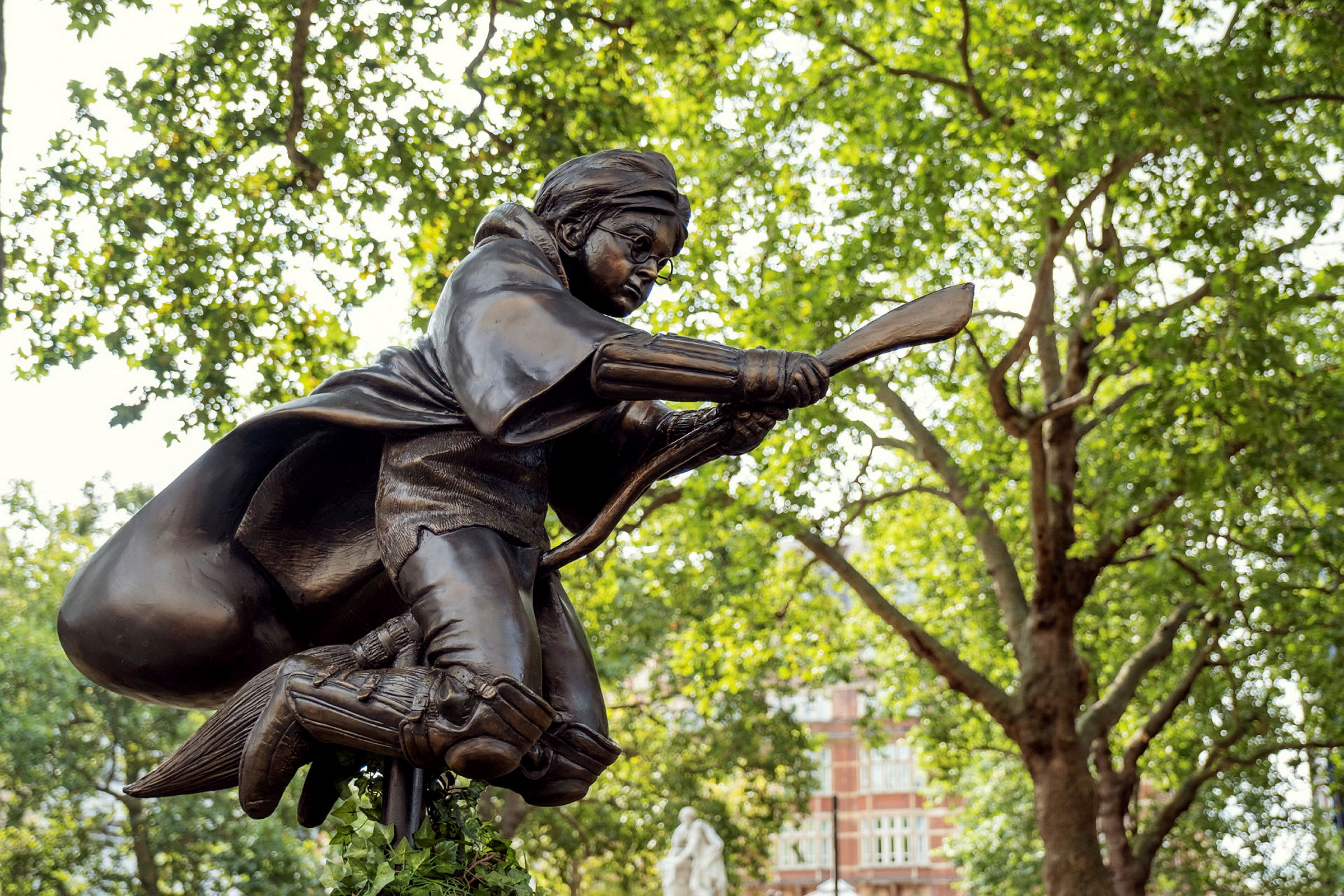 En statue af en figur, der flyver på en kost, omgivet af træer og bygninger. (AI-genereret tekst)