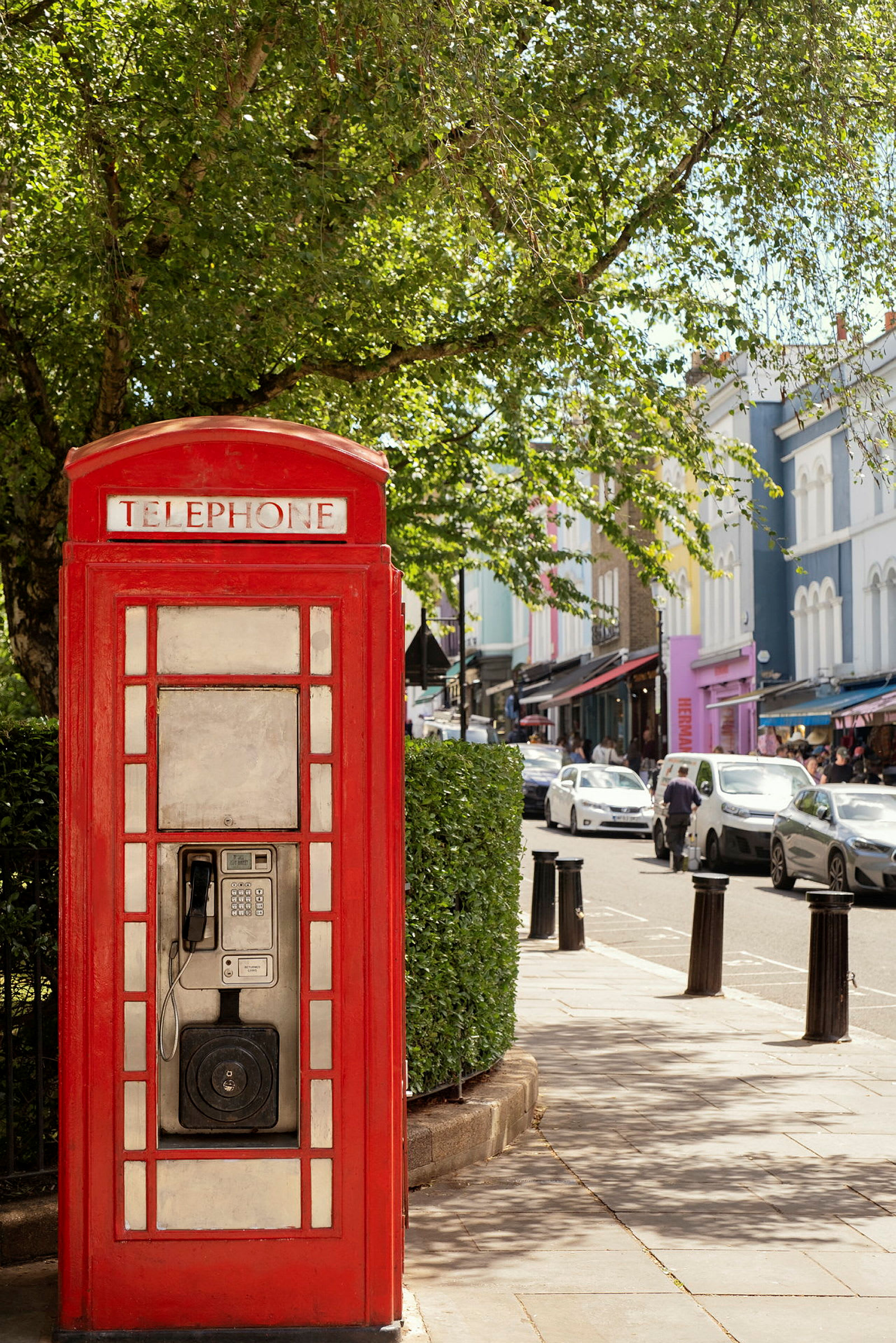 Rød telefonkiosk med grøn bevoksning og by gade i baggrunden. (AI-genereret tekst)