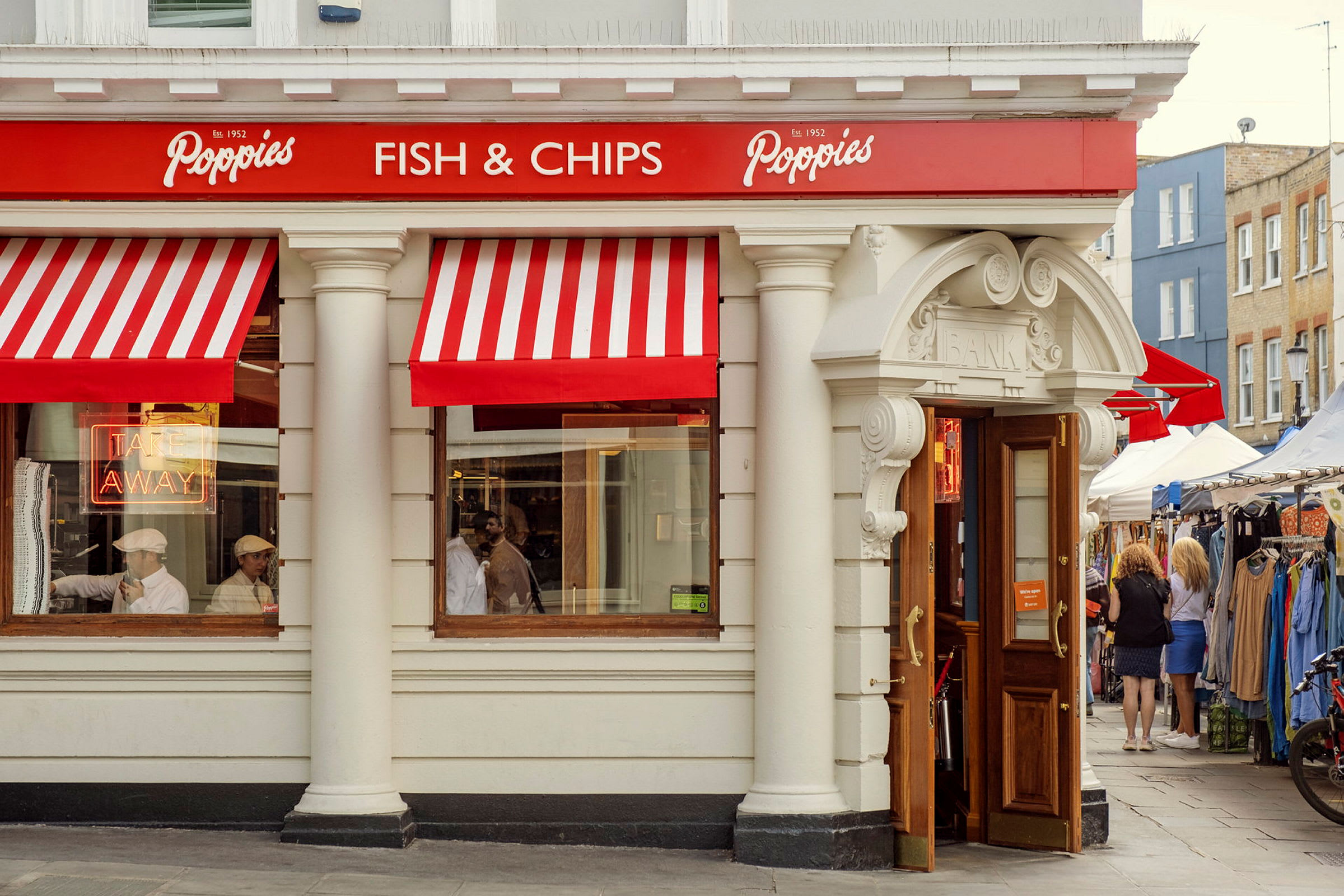 Facaden på en restaurant med røde og hvide markiser, skiltet 'Fish & Chips', og åbne døre. (AI-genereret tekst)