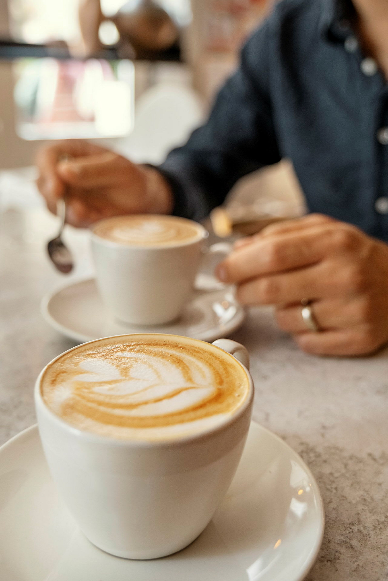 To cappuccino i hvide kopper på et bord, med en hånd der holder en ske. (AI-genereret tekst)