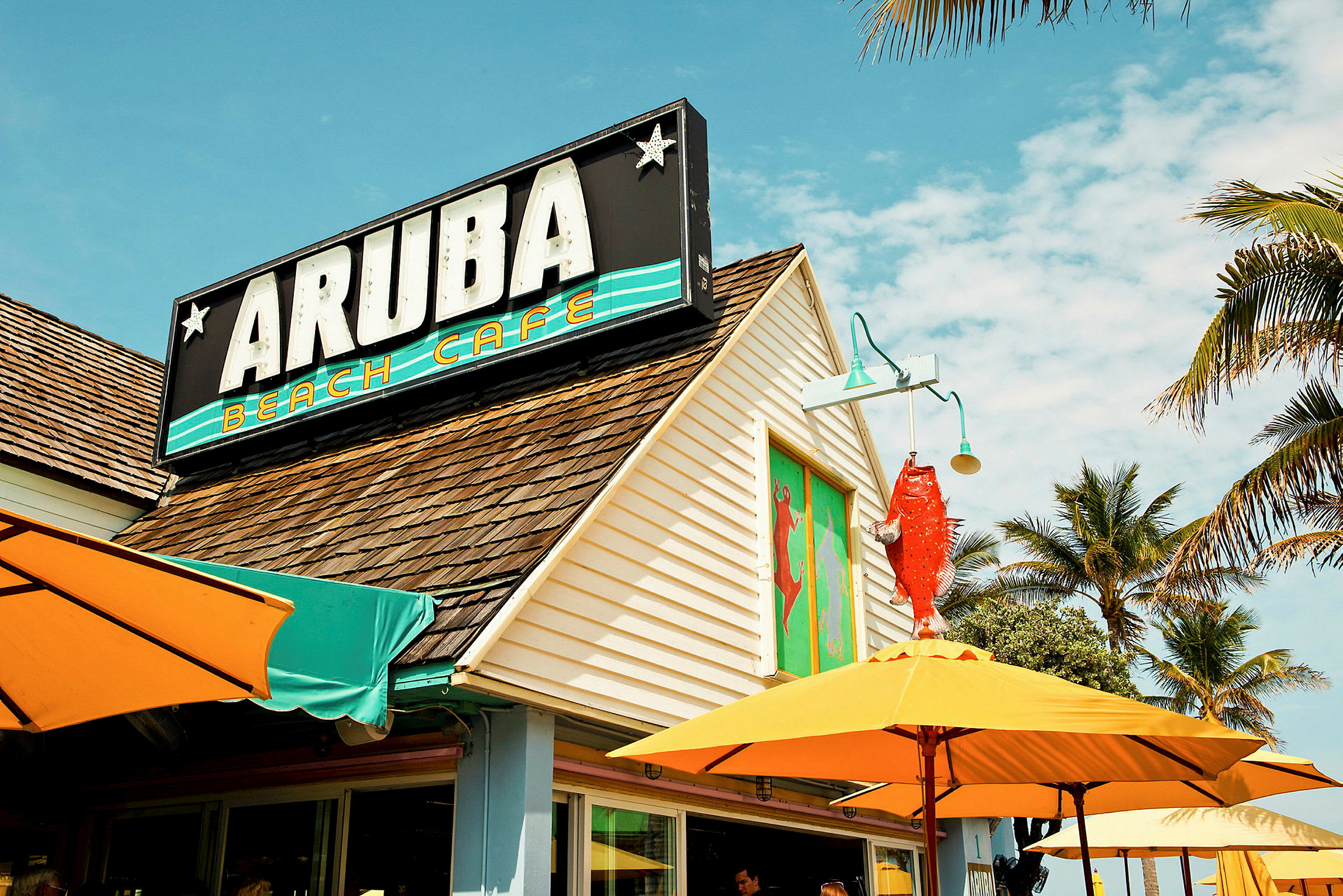 Bygning med et skilt med teksten "Aruba Beach Cafe" og tag med brune tagsten. Farverige parasoller og palmer ses omkring caféen. (AI-genereret tekst)