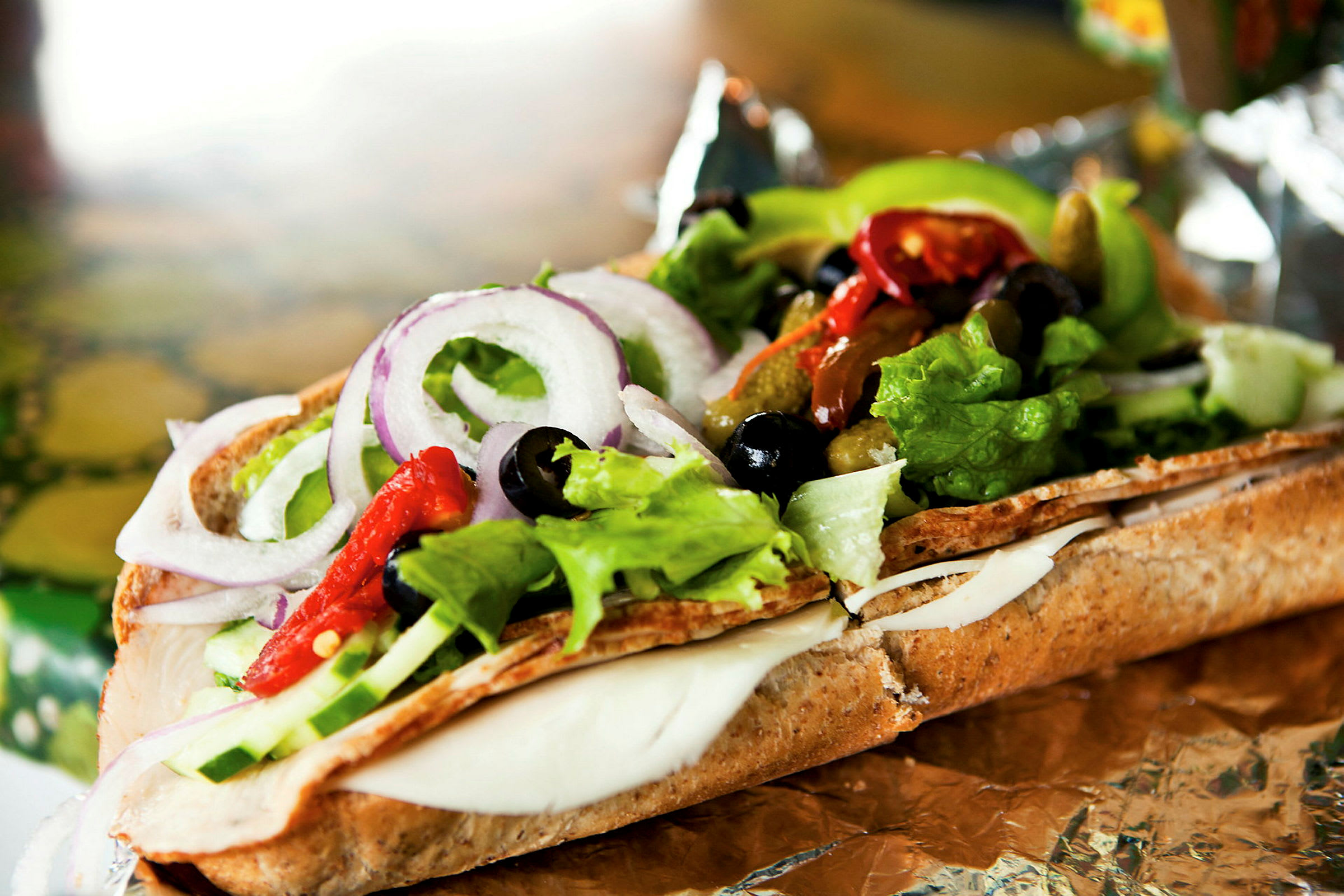 Et nærbillede af en sandwich fyldt med salat, ost, rødløg, oliven og peber på folie. (AI-genereret tekst)