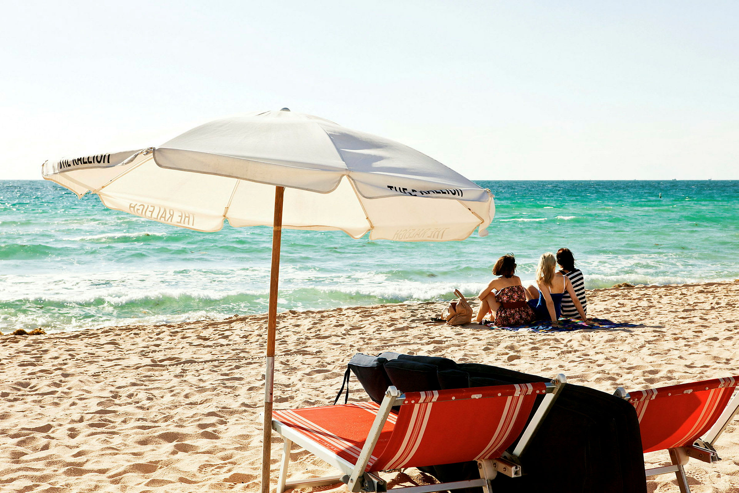 En strand med en parasol og liggestole, samt tre personer, der sidder og nyder udsigten over havet. (AI-genereret tekst)