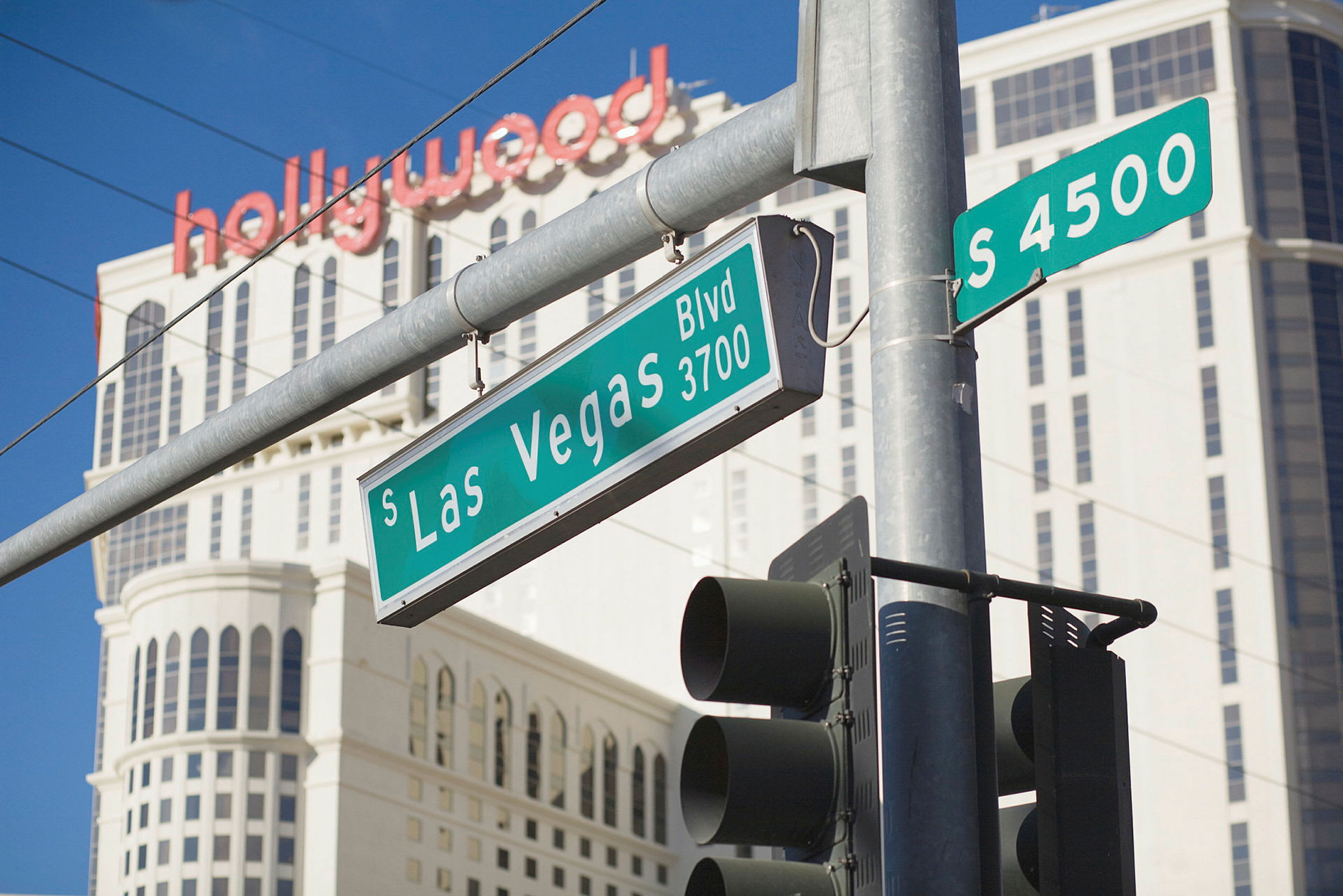 Fodgængerfelt med trafiksignal og vejskilte ved South Las Vegas Boulevard, med en høj bygning og teksten "hollywood" i baggrunden. (AI-genereret tekst)