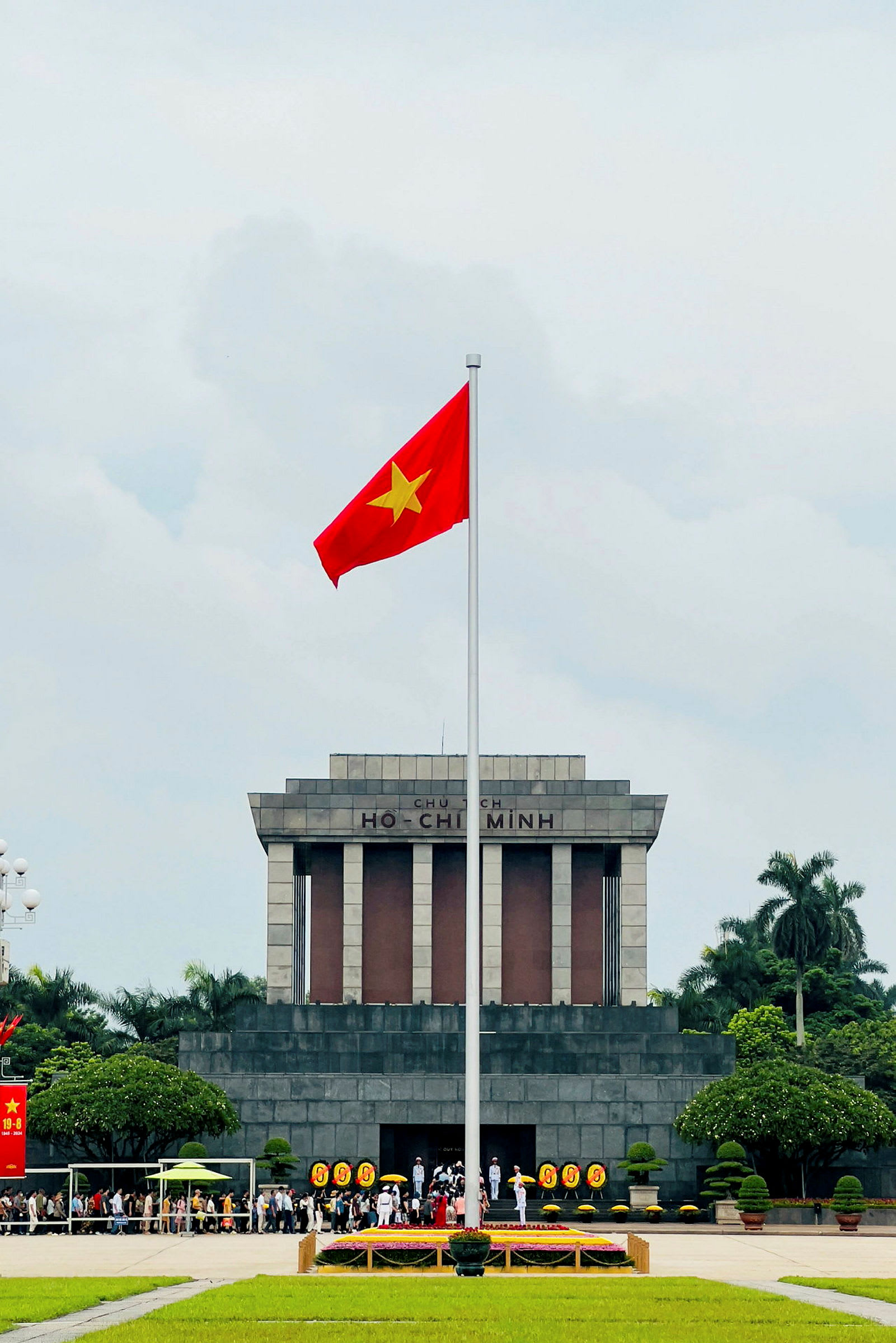 Et stort mausoleum med teksten 'Ho Chi Minh' på facaden, foran hvilket det vietnamesiske flag er rejst på en flagstang. Folk samles foran bygningen, som er omgivet af træer og græsplæner. (AI-genereret tekst)